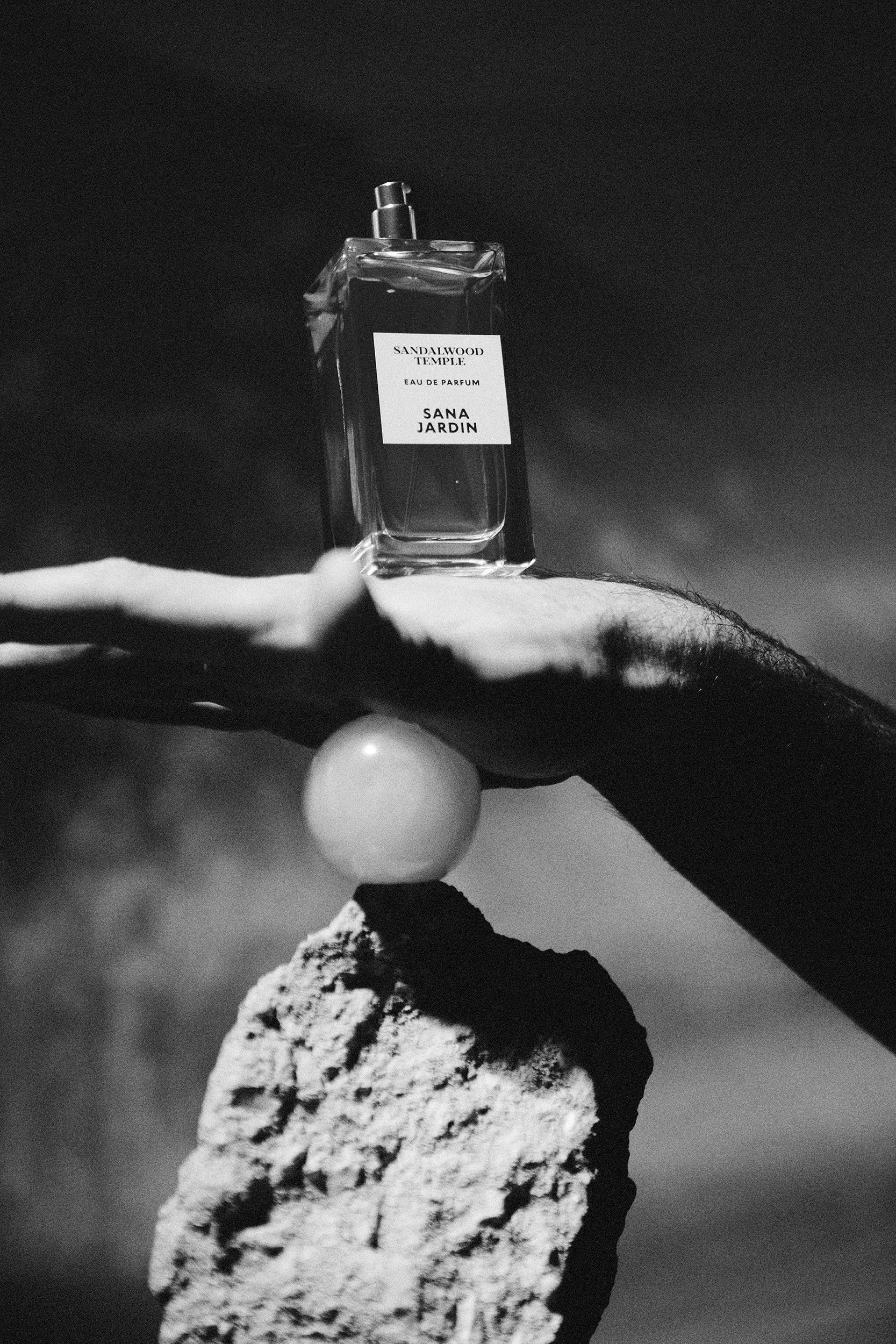Black and white photo of a hand balancing on a rock with a spherical object, supporting a perfume bottle labeled 'Sandalwood Temple,' 'Eau de Parfum,' 'Sana Jardin'.