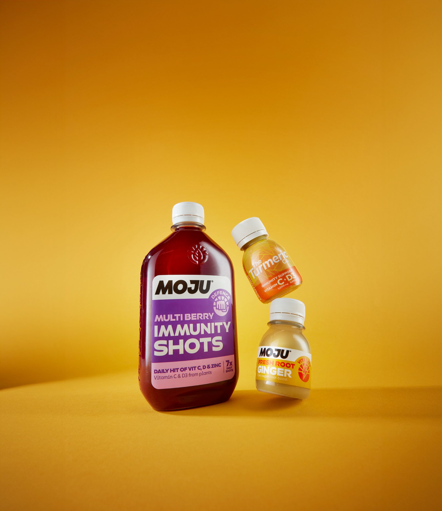 Three bottles of health supplements on a yellow background, including multi berry immunity shots, turmeric, and ginger.