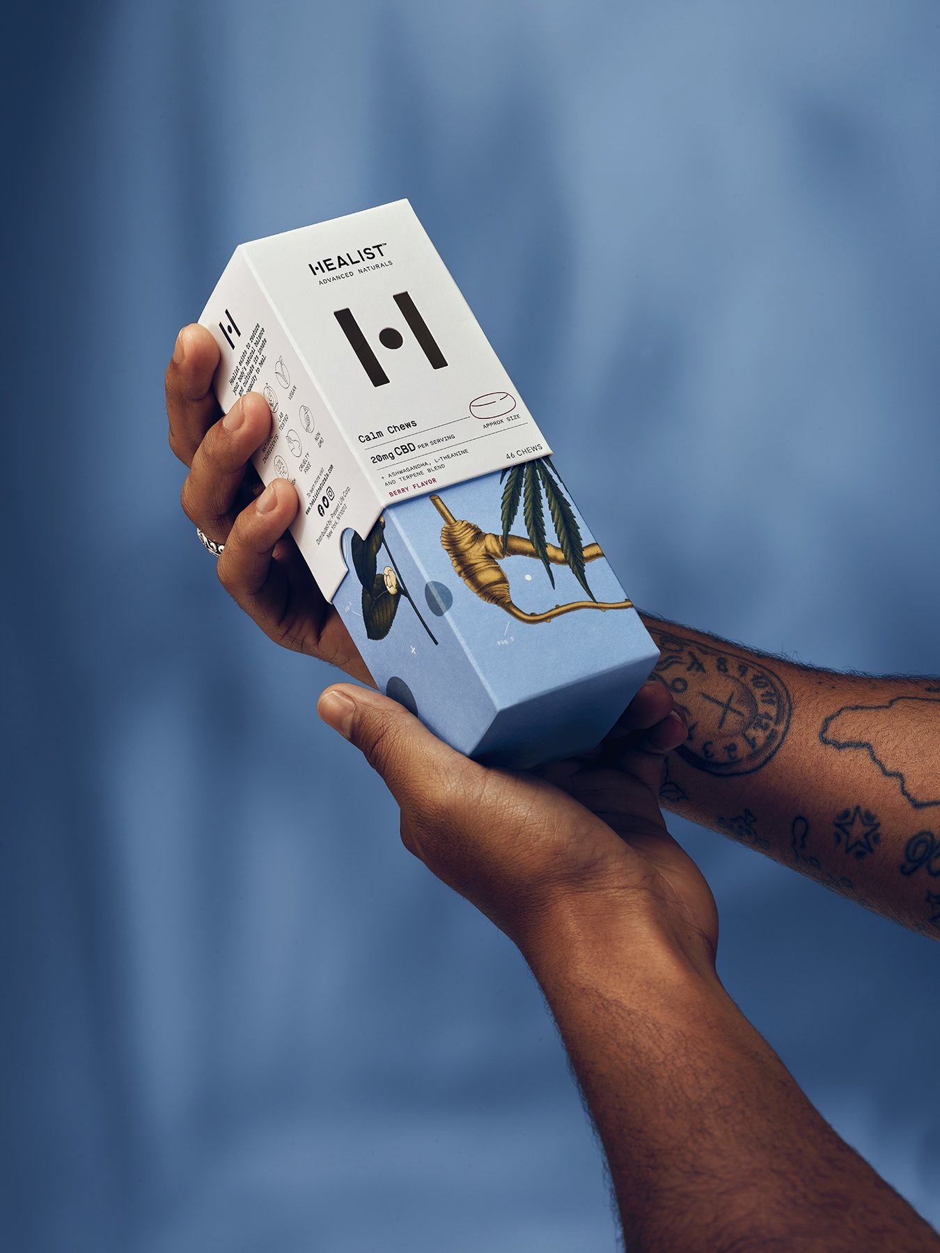 A person holding a box of HEALIST CBD chews, featuring a design with a marijuana leaf, a bee, and colorful artwork on a blue background.