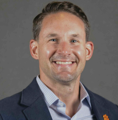 A smiling man in a suit jacket and collared shirt against a gray background.