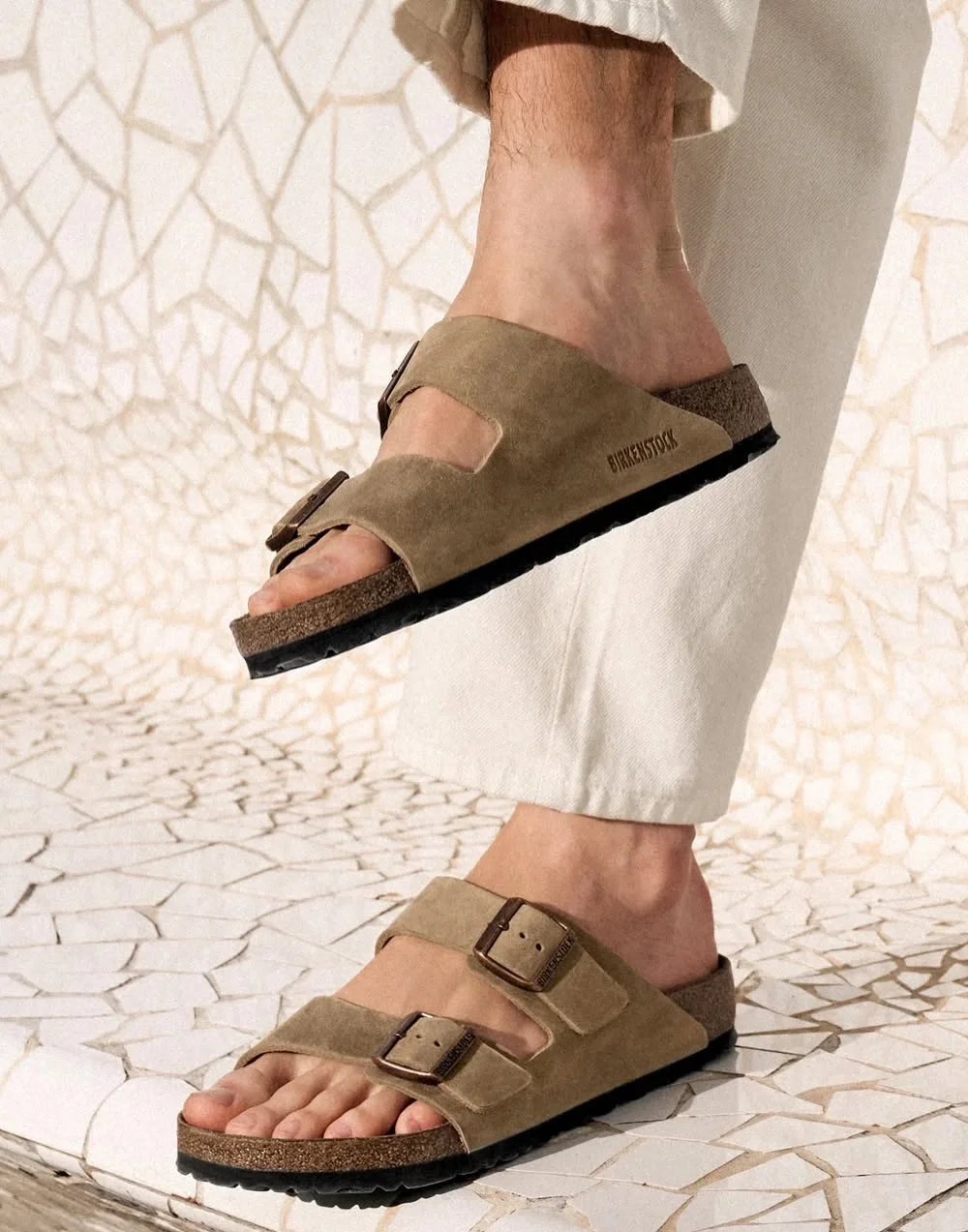Close-up of a person wearing beige Birkenstock sandals with adjustable straps, standing on a textured, cracked white surface.