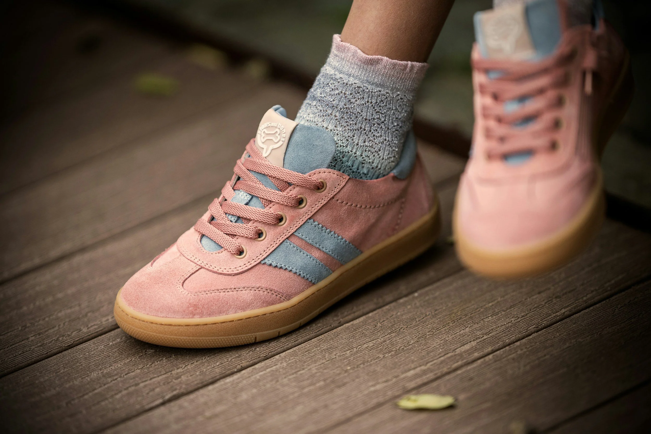 Close-up of a person wearing pink and blue sneakers with beige soles and gray patterned socks, standing on a wooden surface outdoors.