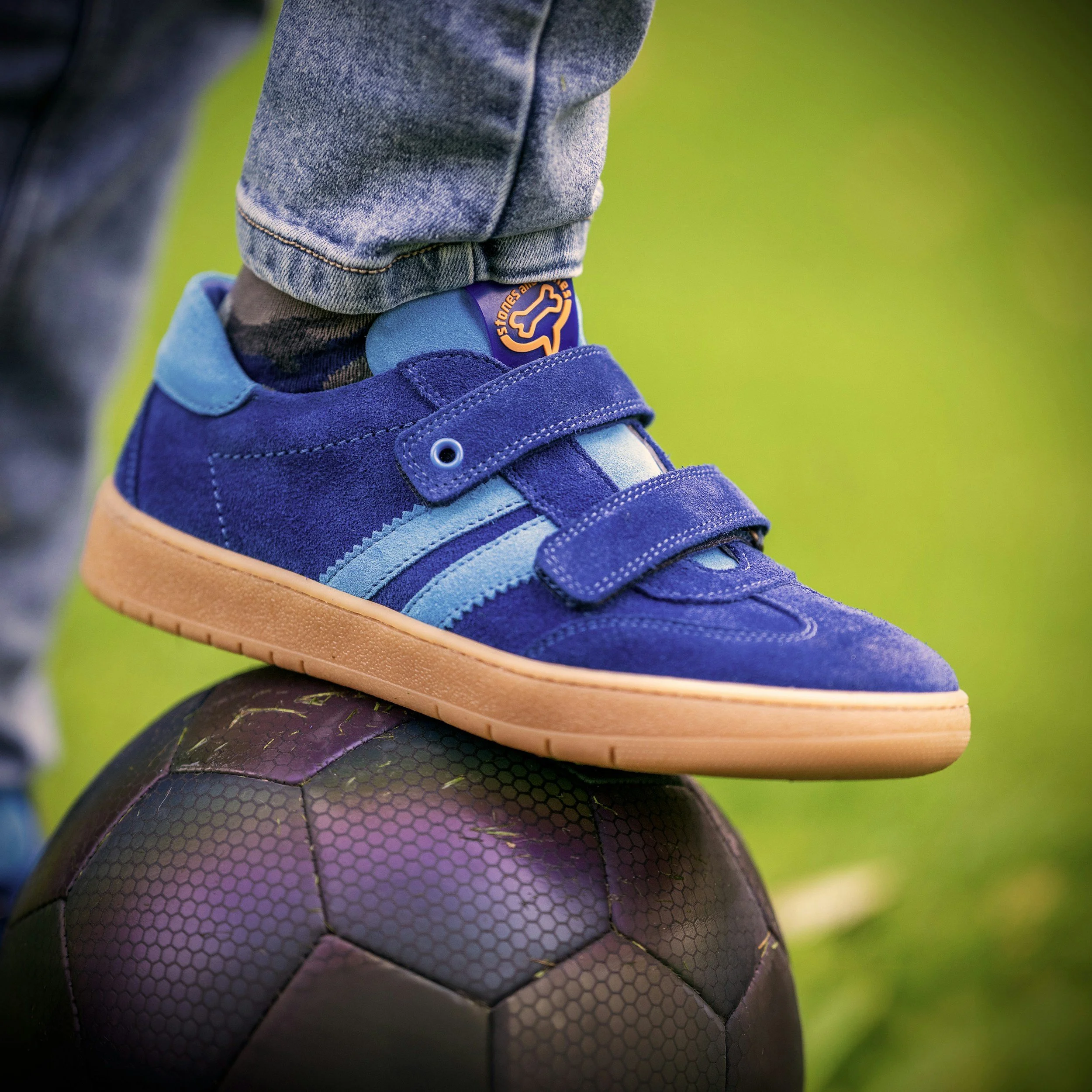 Close-up of a person's foot wearing a blue sneaker with orange accents, resting on a purple and black textured soccer ball, set against a blurry green background.