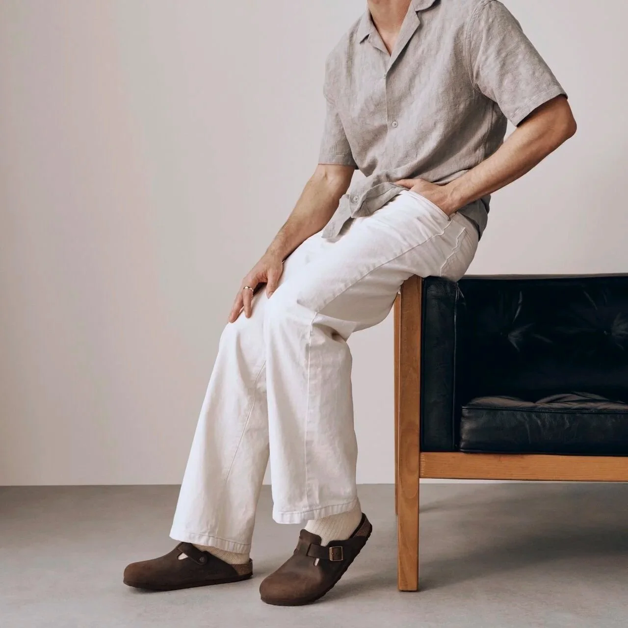 A person sitting on the edge of a black leather and wood bench with one hand resting on their lap and the other supporting them, wearing a light gray short-sleeve button-up shirt, white pants, and brown shoes, against a plain white wall background.