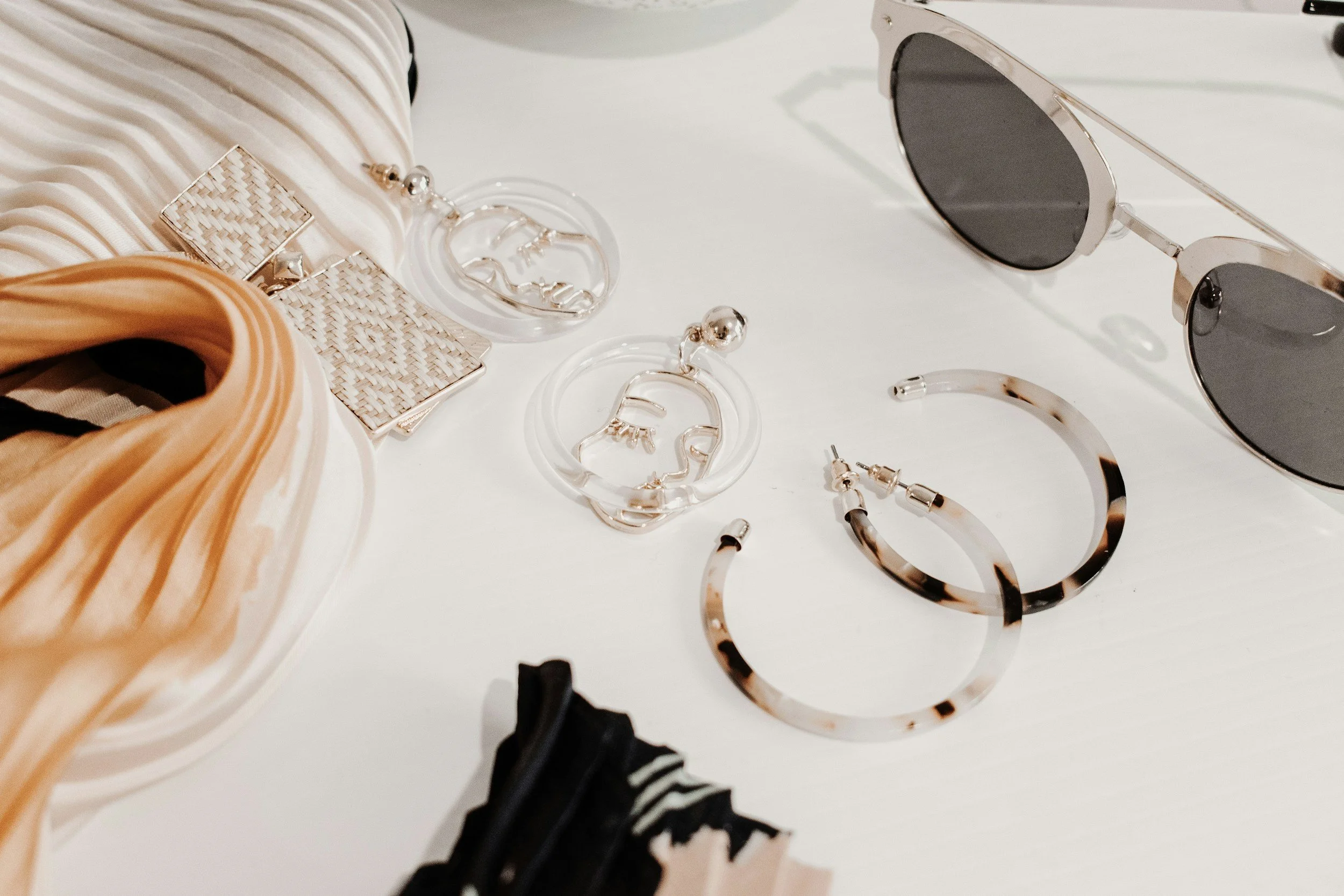 A collection of fashion accessories including sunglasses, earrings, a bracelet, and a makeup palette on a white surface.
