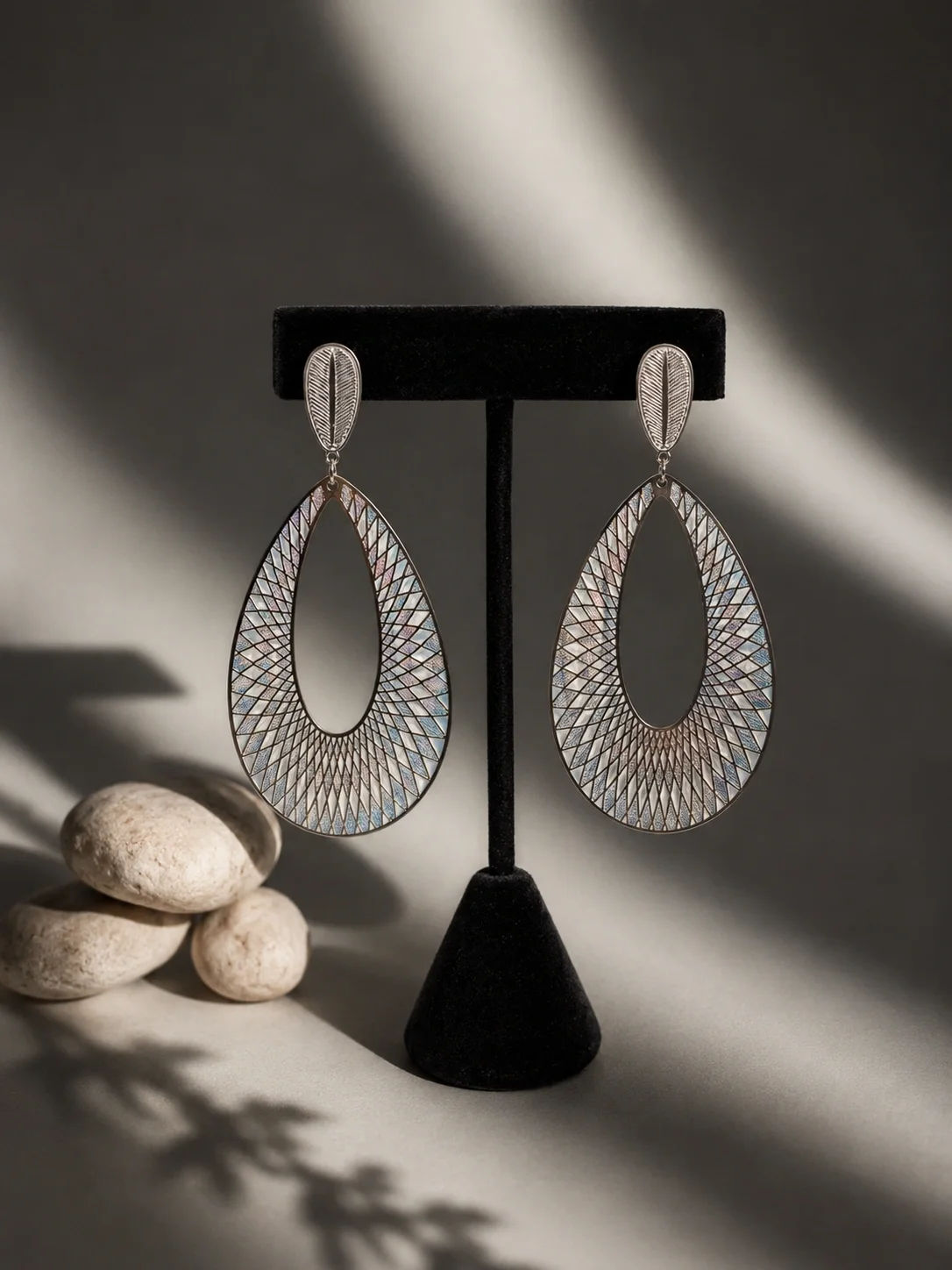 Silver teardrop-shaped earrings with intricate geometric pattern displayed on a black stand, with three stacked white rocks and soft shadowed background.