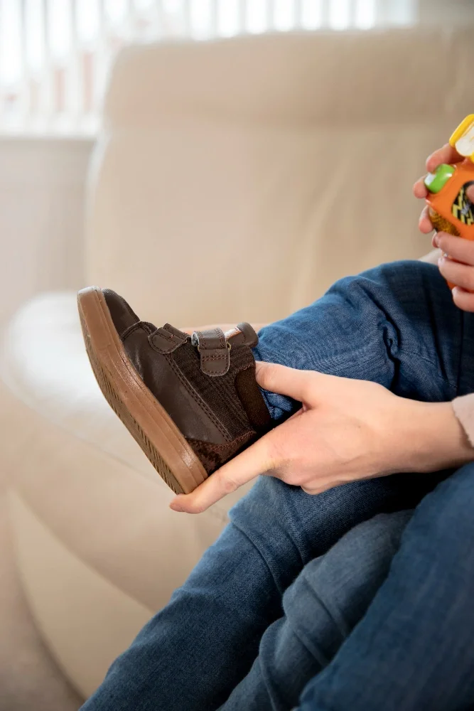 Child sitting on a beige couch, holding a brown shoe with velcro straps, while holding a yellow and green toy in one hand.