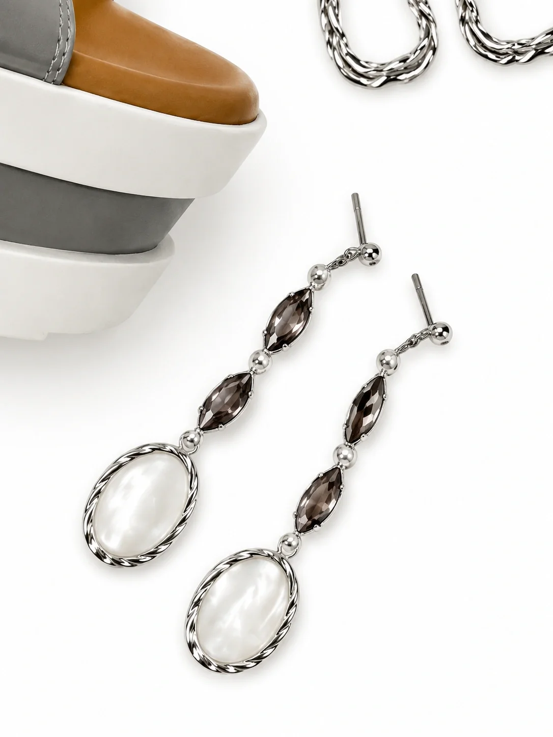 Pair of dangling earrings with oval chain links, dark gemstones, and white oval stones, placed on a white surface near part of a jewelry box and a silver chain