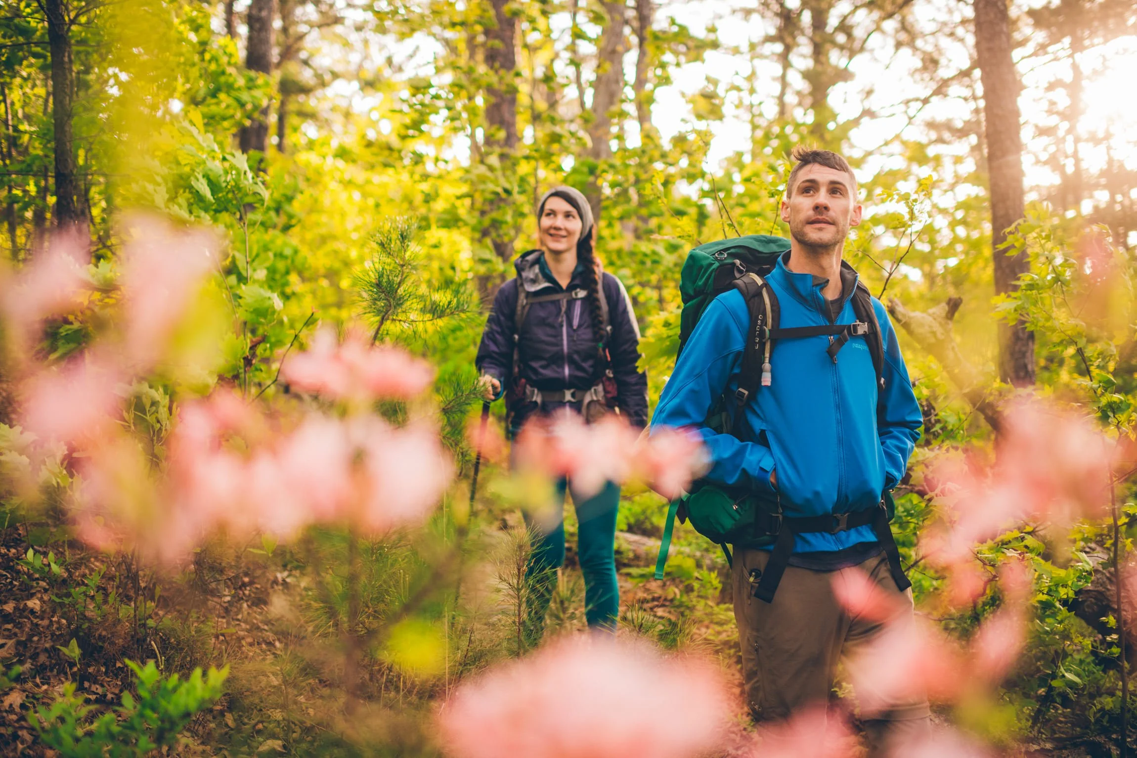 Two hikers, a man and a woman, walking through a lush green forest during daytime, with sunlight filtering through the trees.