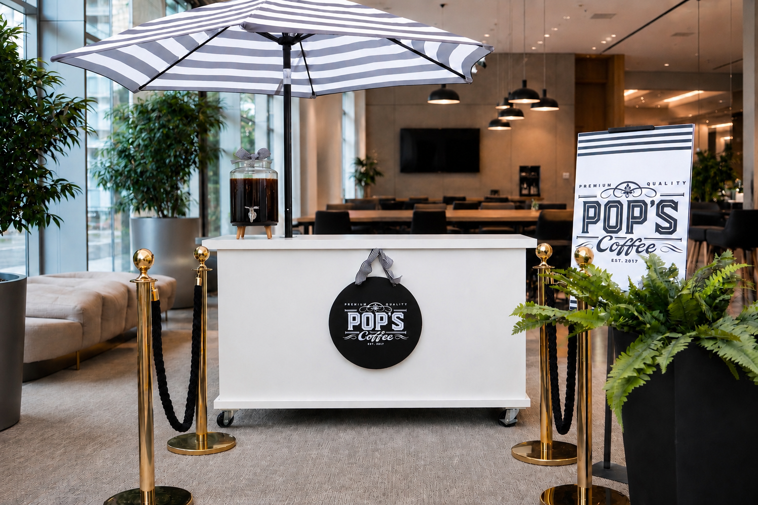 A coffee stand named POP'S Coffee with a black and white striped umbrella, black sign, and gold stanchions in a modern indoor setting with plants and seating.
