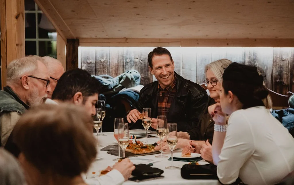 En gruppe voksne og unge som spiser og prater rundt et bord i et rom med trepanel på veggene.