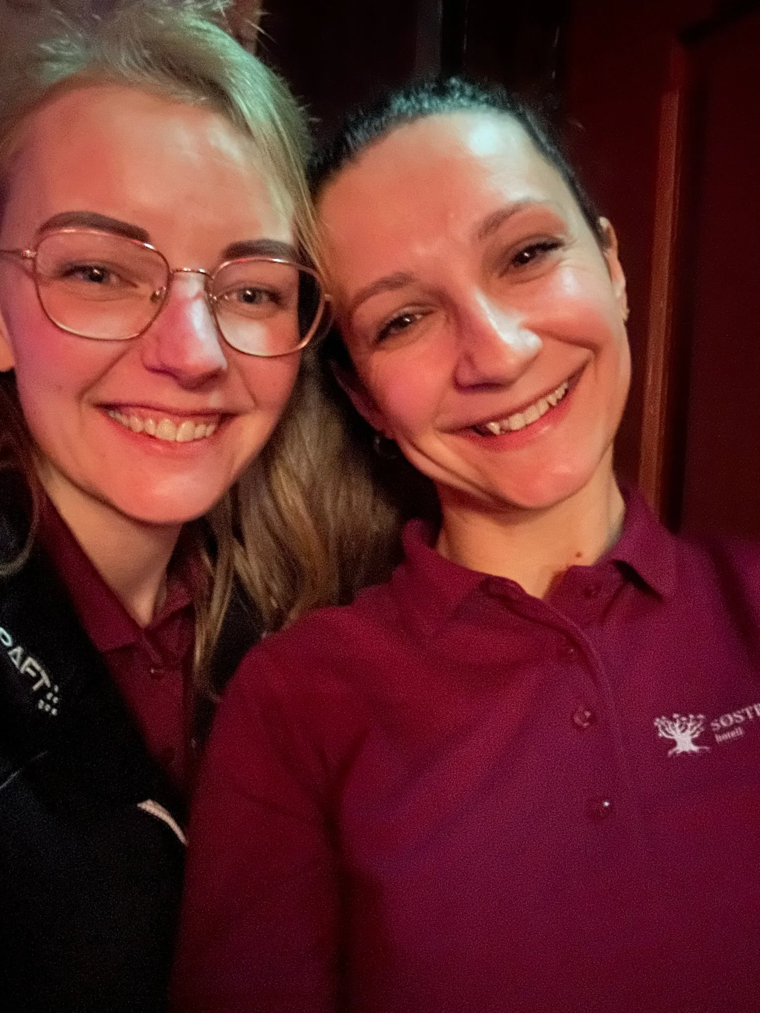 To kvinner smiler og tar en selfie, den ene har briller og blondt hår, den andre har mørkt hår og er kledd i rød uniform med logo.
