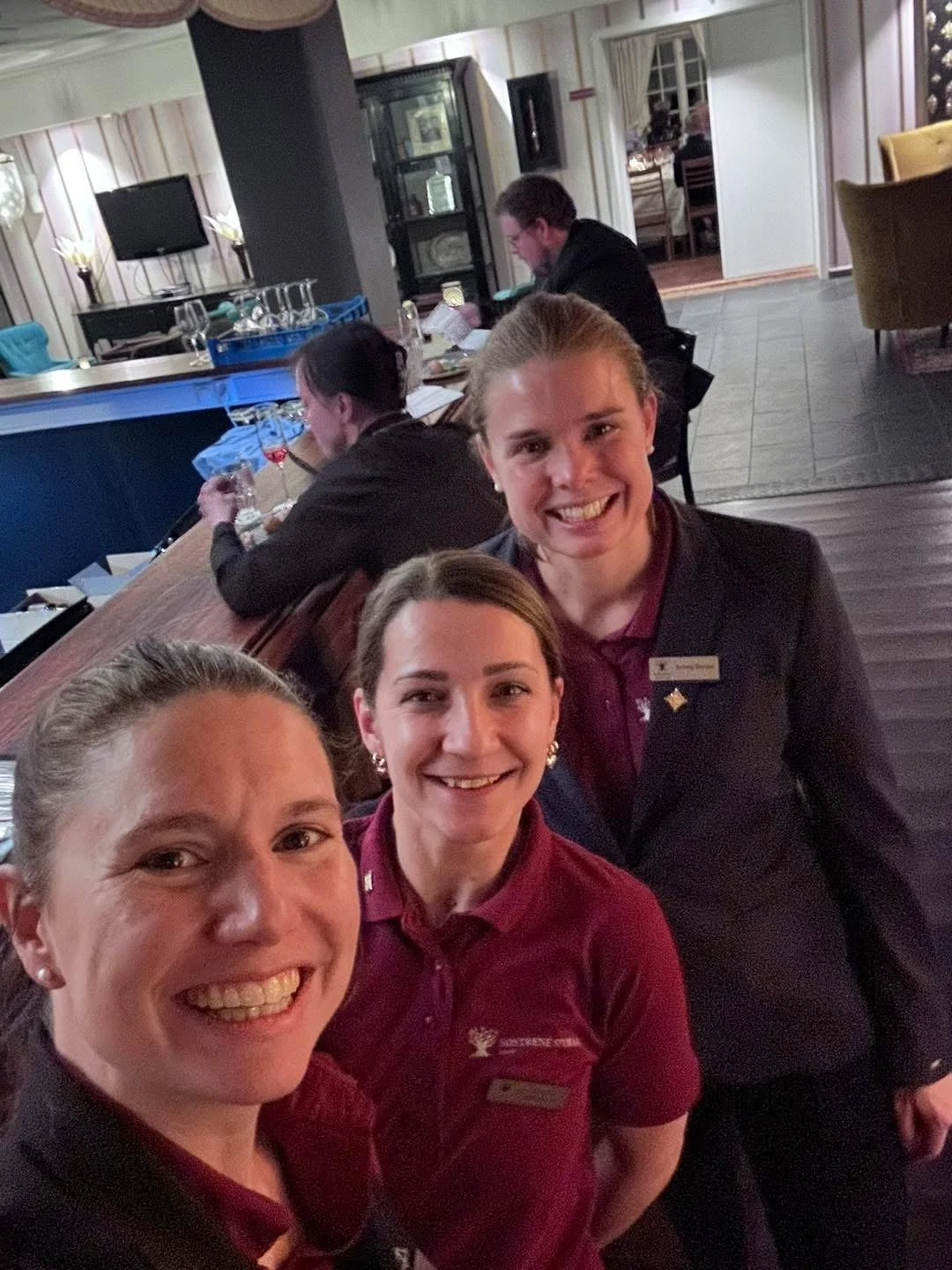 Tre smilende kvinner og en mann i uniformer tar selfie i en restaurant. Andre personer i bakgrunnen som spiser og leser meny.