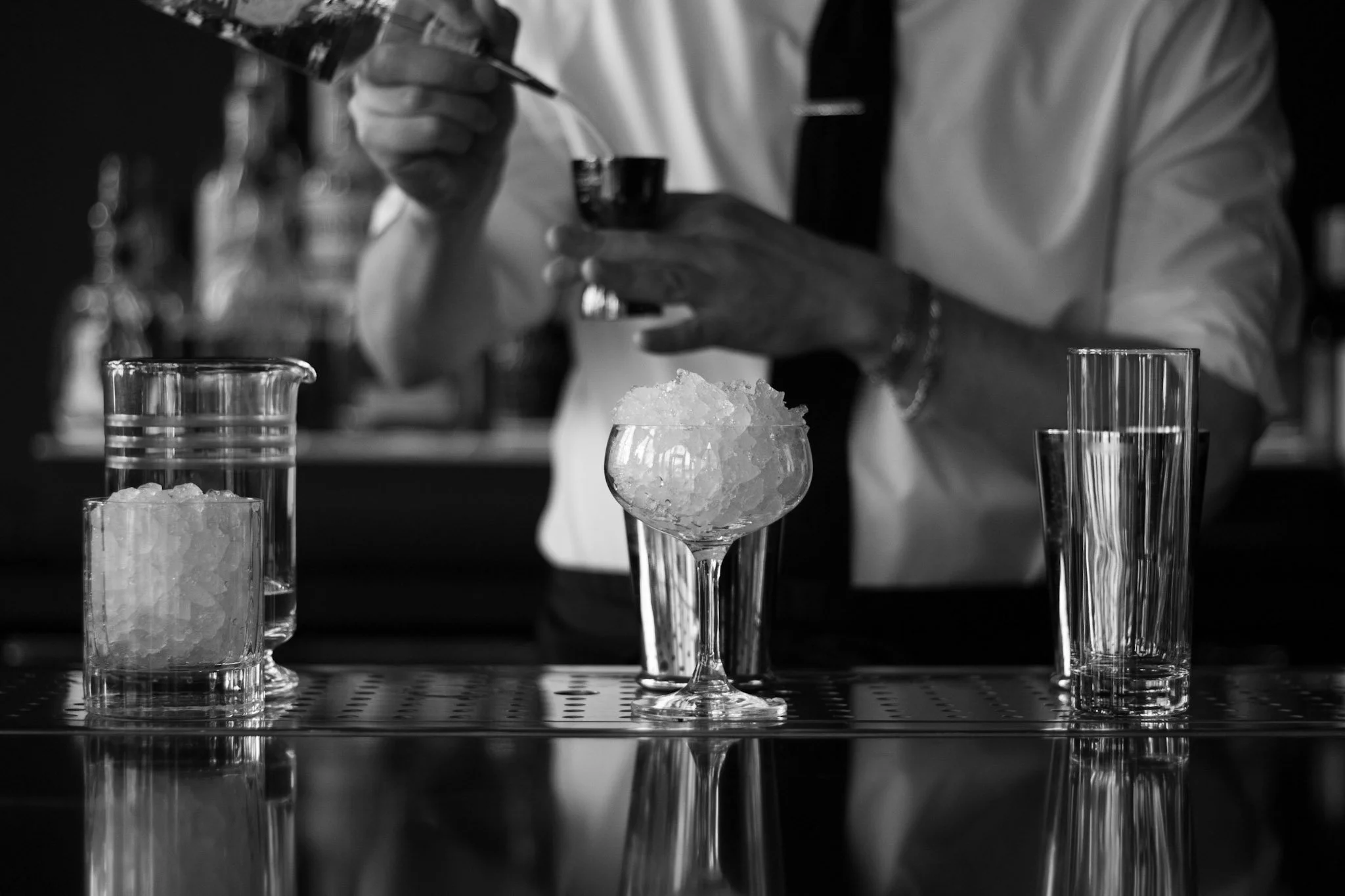 A bartender in a white shirt and dark apron pours a drink into a glass with crushed ice, surrounded by various glassware on a bar counter.