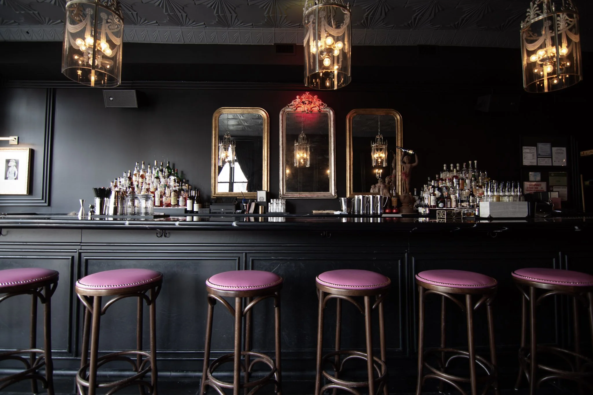 A dimly lit bar with black walls, barstools with pink seats, gold-framed mirrors, and a bar counter with various liquor bottles and bar accessories, illuminated by elegant chandeliers.