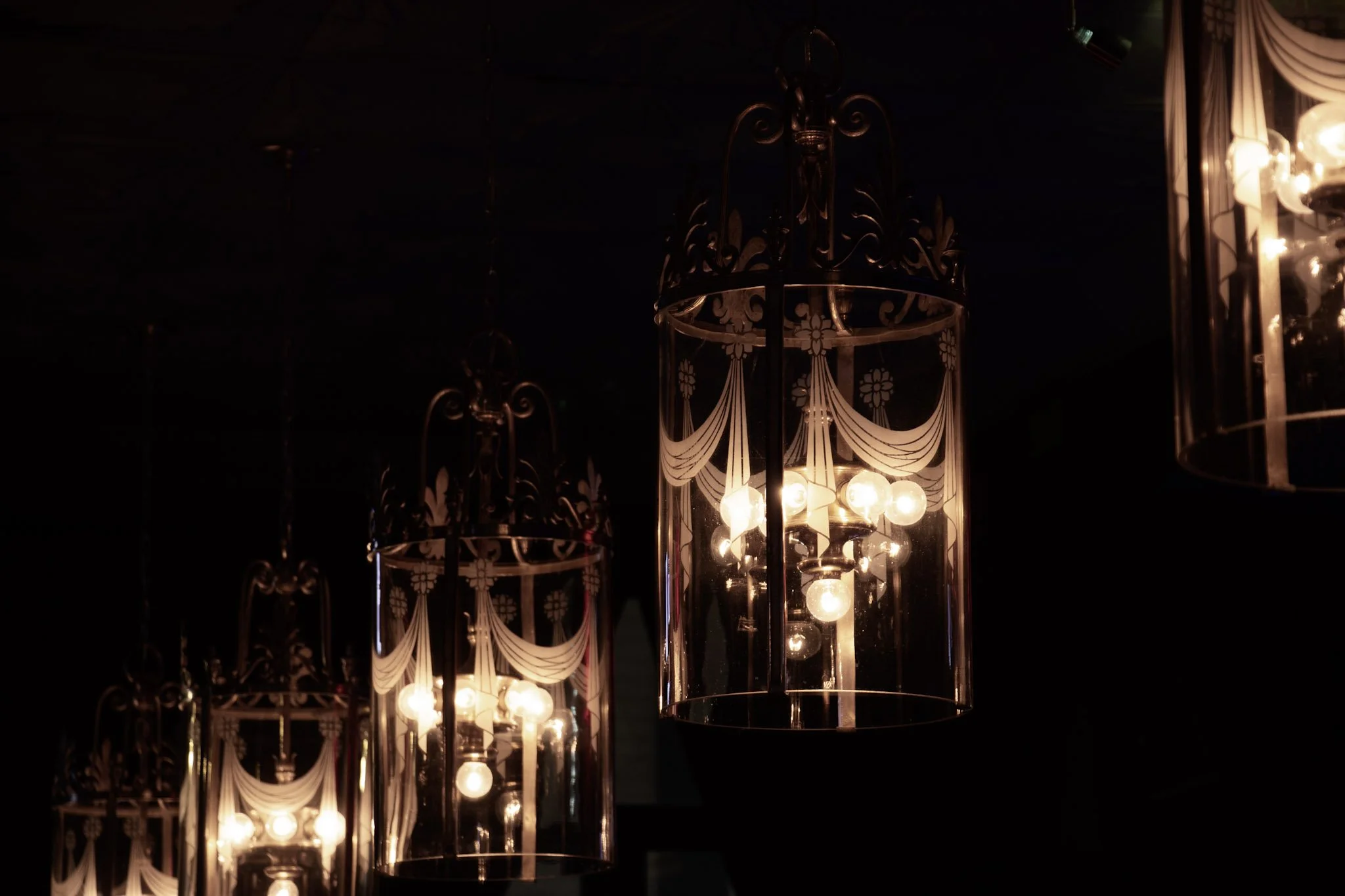 Multiple ornate glass chandeliers with bulbs inside, hanging in a dark room, illuminated.