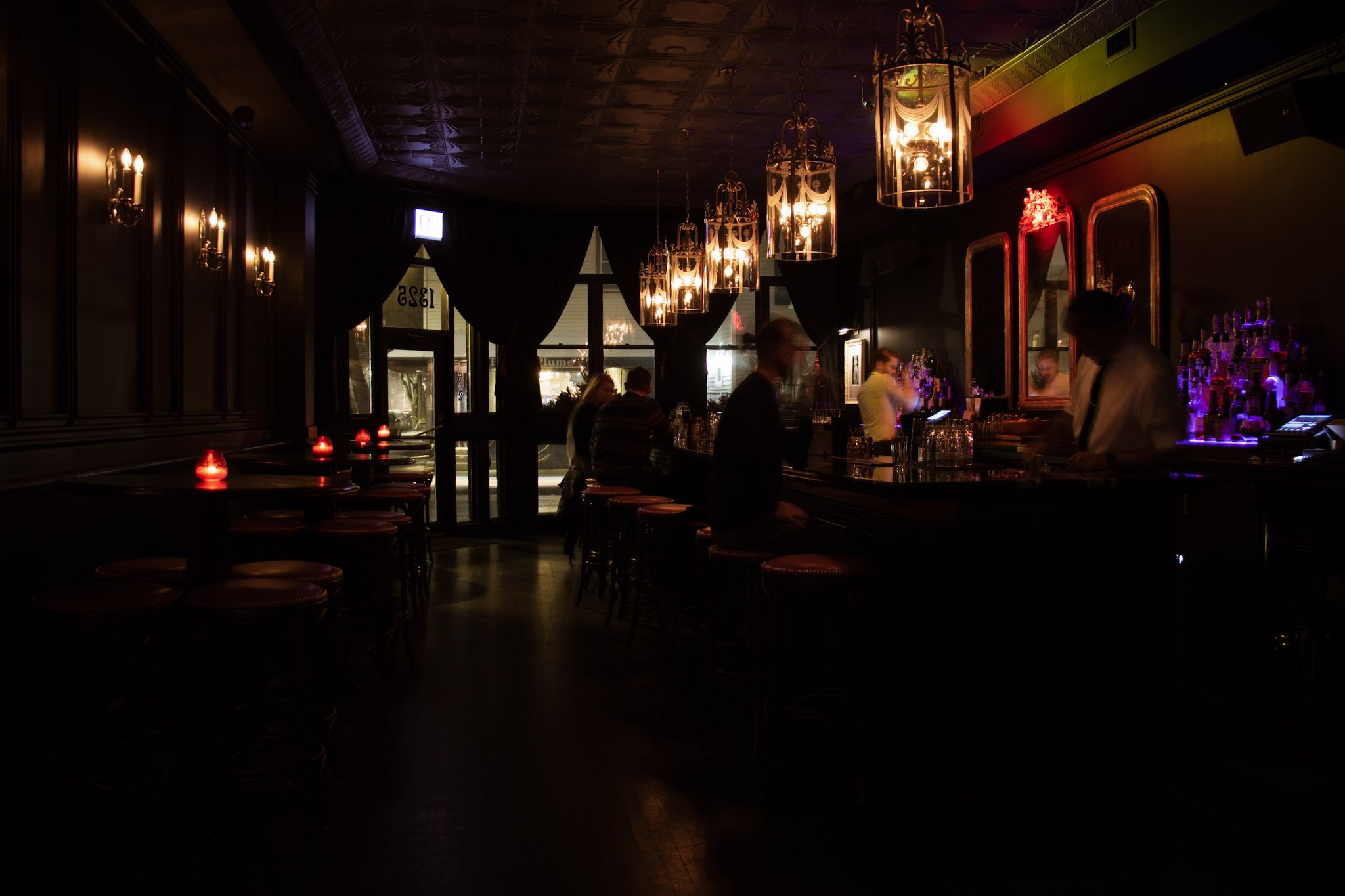 Interior of a dimly lit bar with hanging chandeliers, a bar counter with bottles and glasses, and a few people sitting and standing.