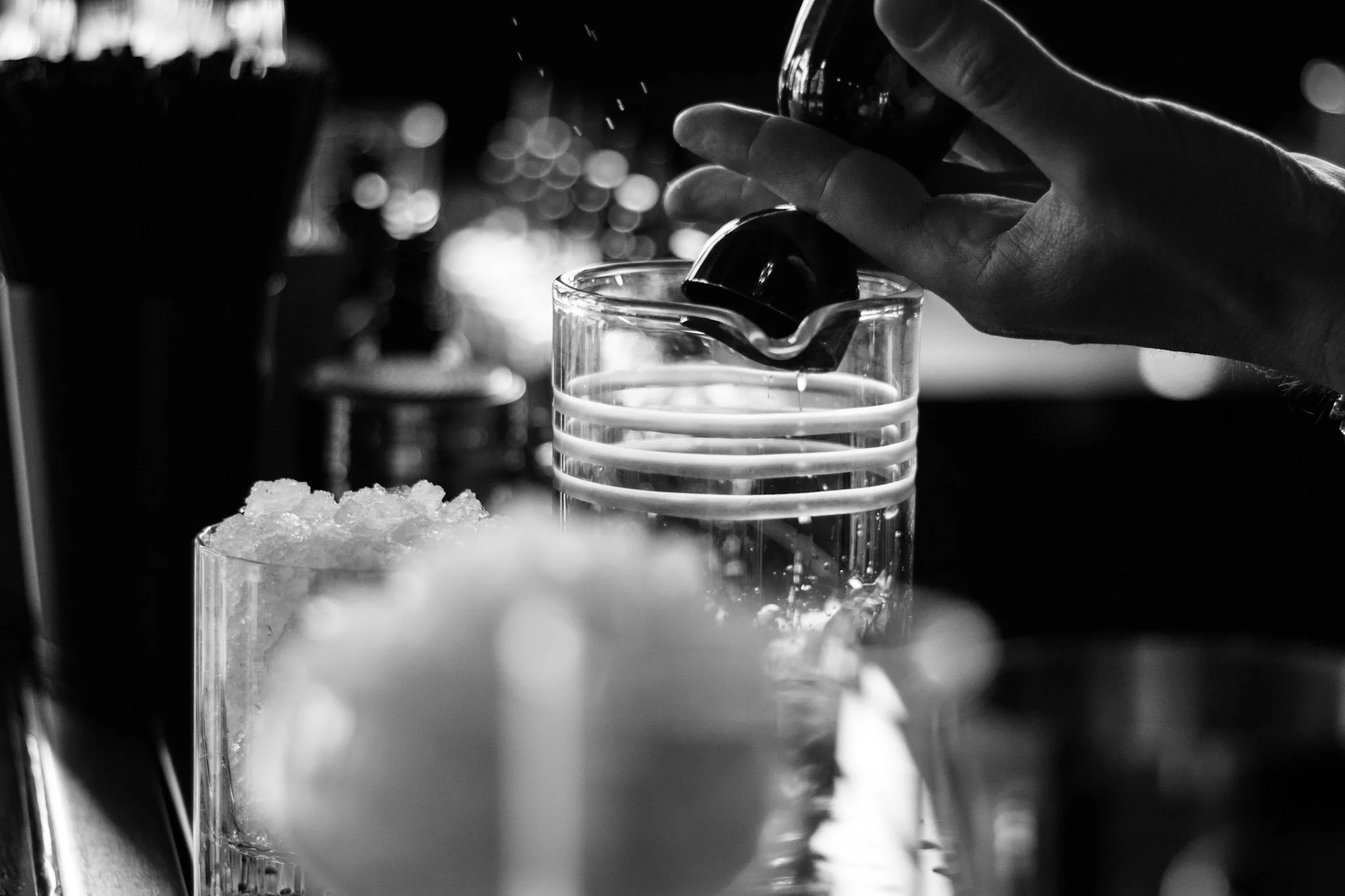 A close-up black and white photo of a hand pouring a dark liquid into a glass, with ice cubes and a blurred background of bottles.