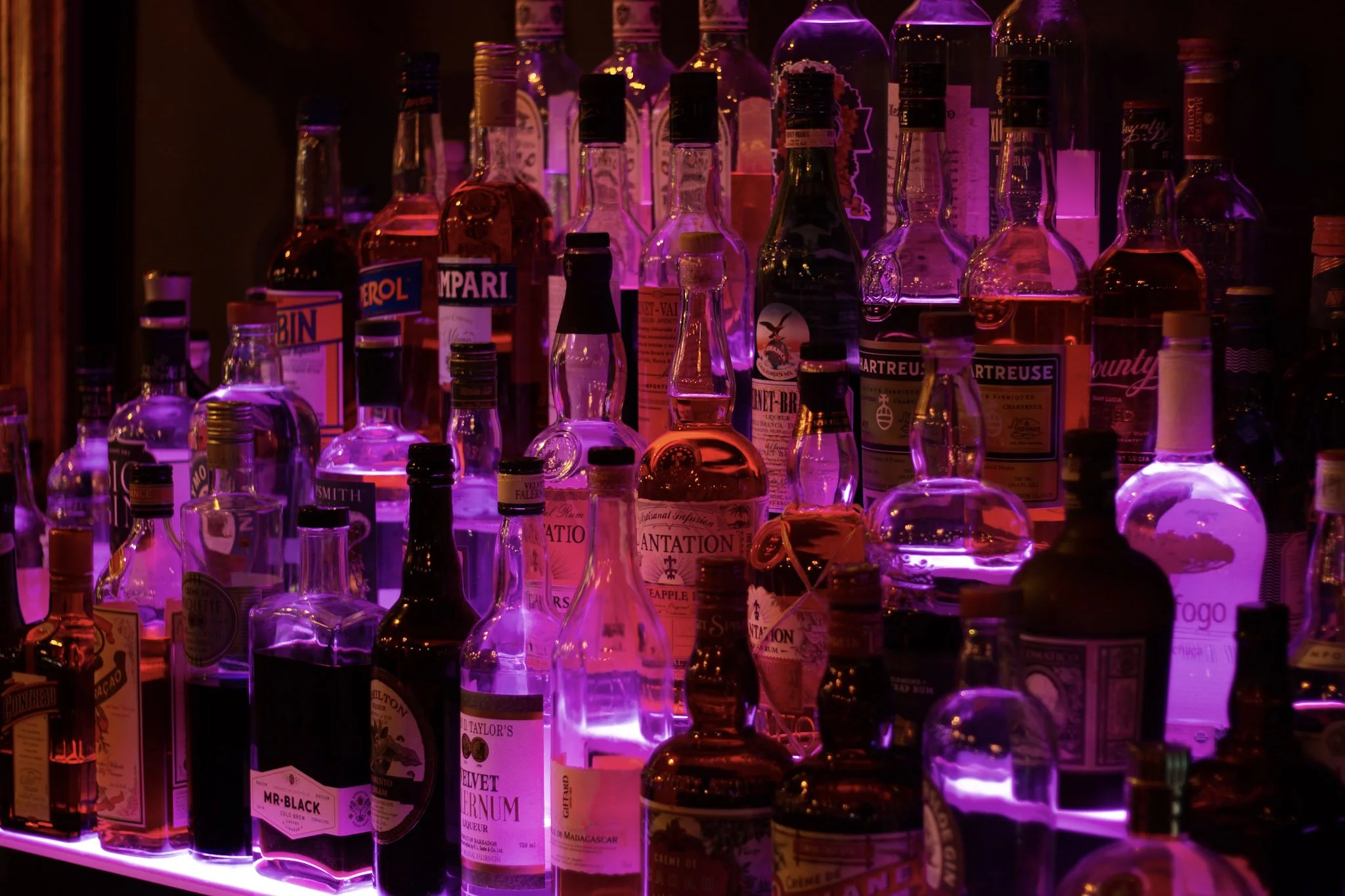 A collection of various liquor bottles on a shelf illuminated by purple lighting.
