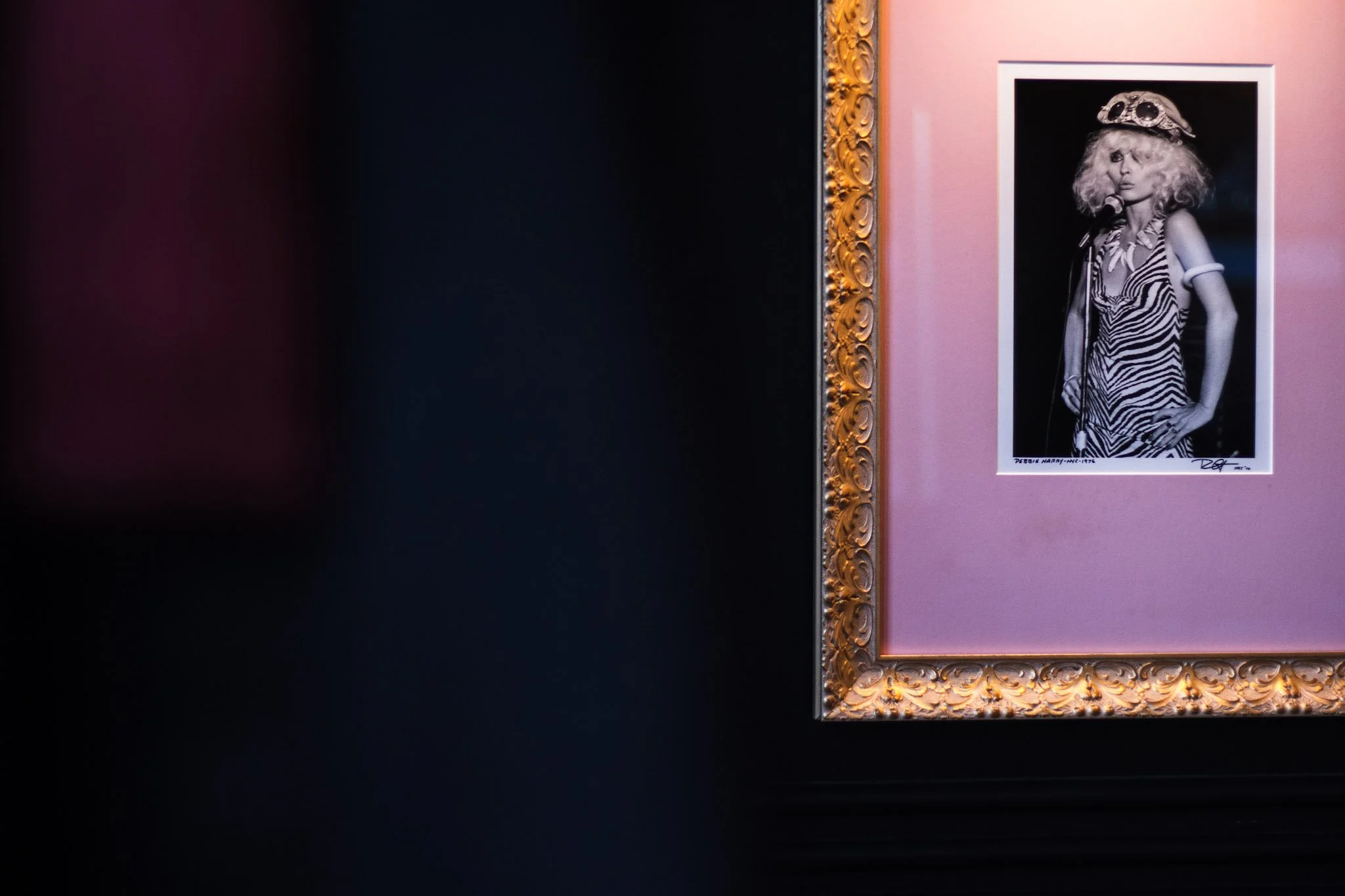 A black and white portrait photograph of a woman with curly hair, wearing a striped dress and a decorative hat. The photo is framed with an ornate gold frame and is displayed on a pink wall.