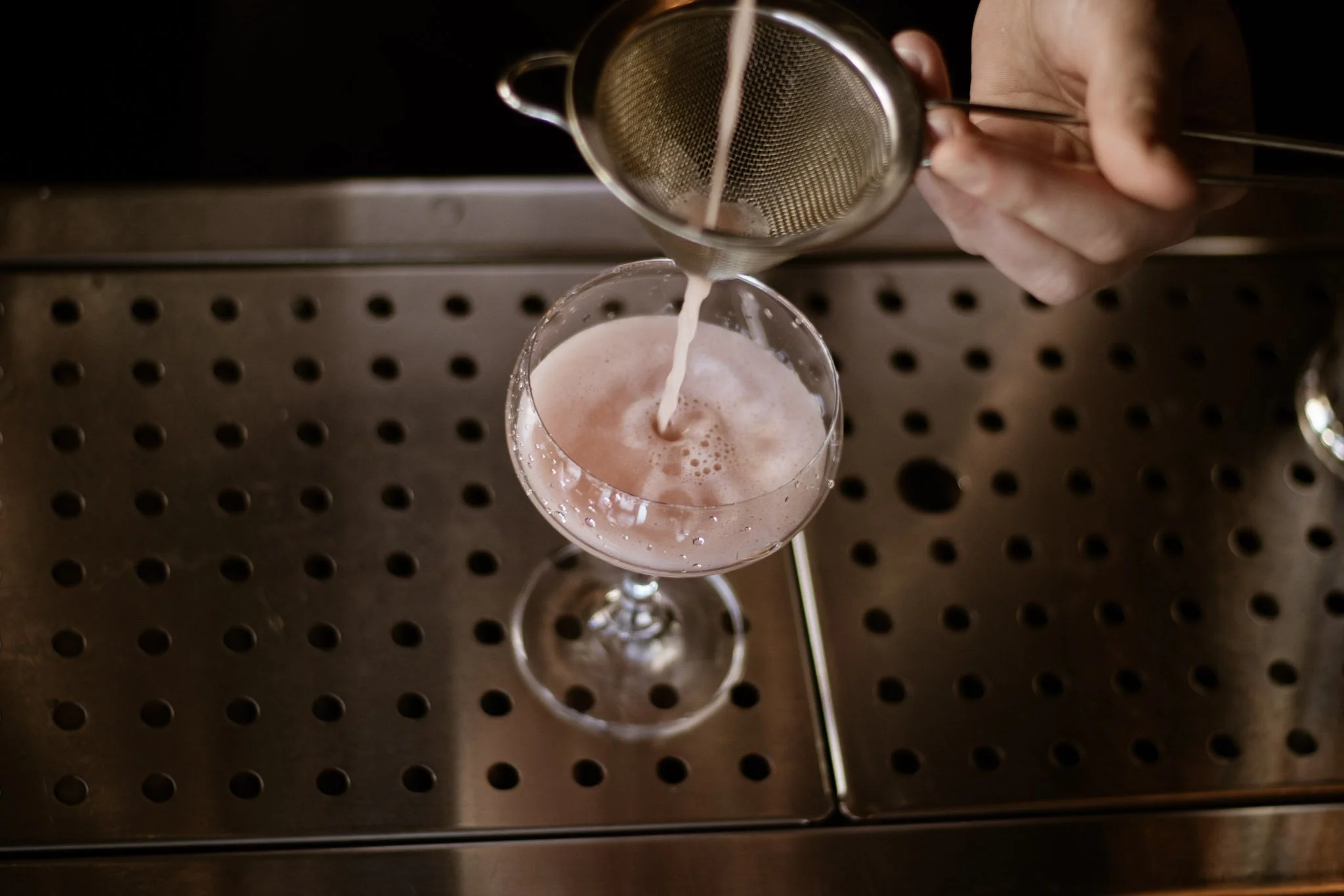 A hand holding a metal strainer pouring frothy pink liquid into a glass wine goblet on a metallic surface.