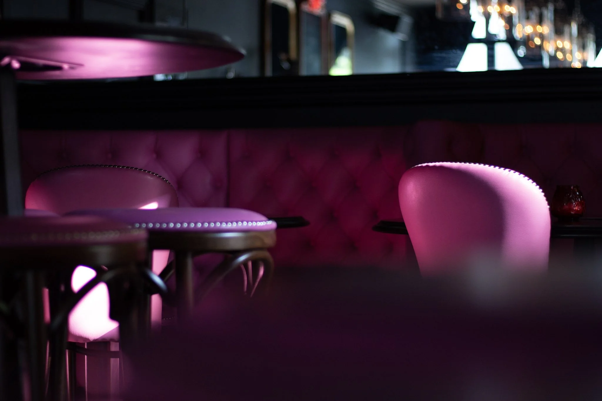 A dimly lit restaurant or bar with pink upholstered seating, round wooden tables, and a bar area with bottles and lights in the background.