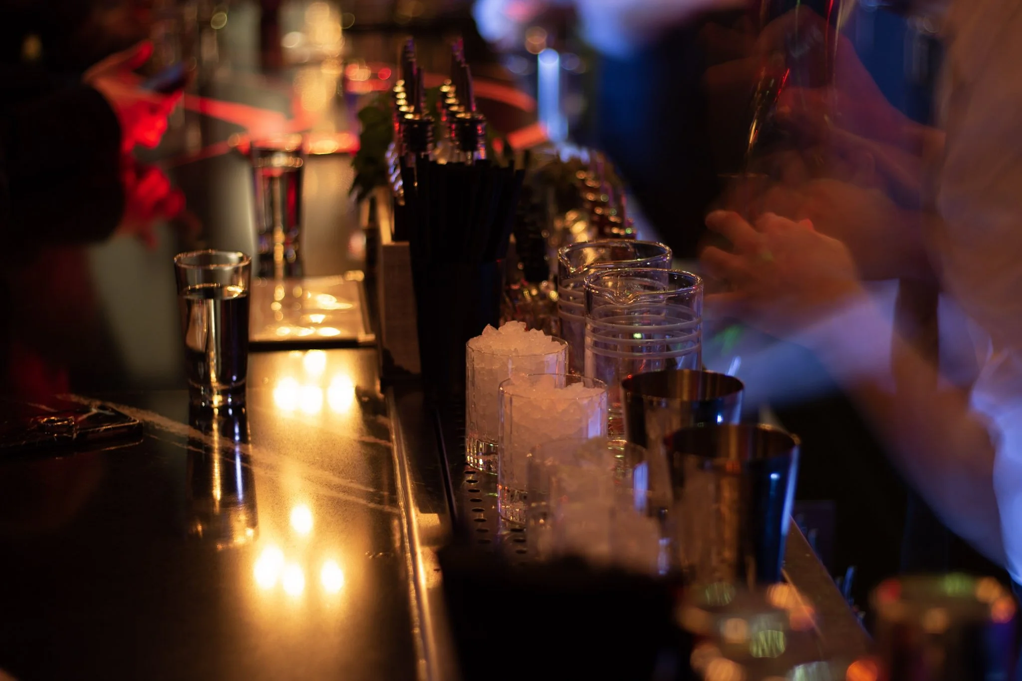 A dimly lit bar with colorful neon lights, glasses, and bottles on the counter, and ice in small containers.