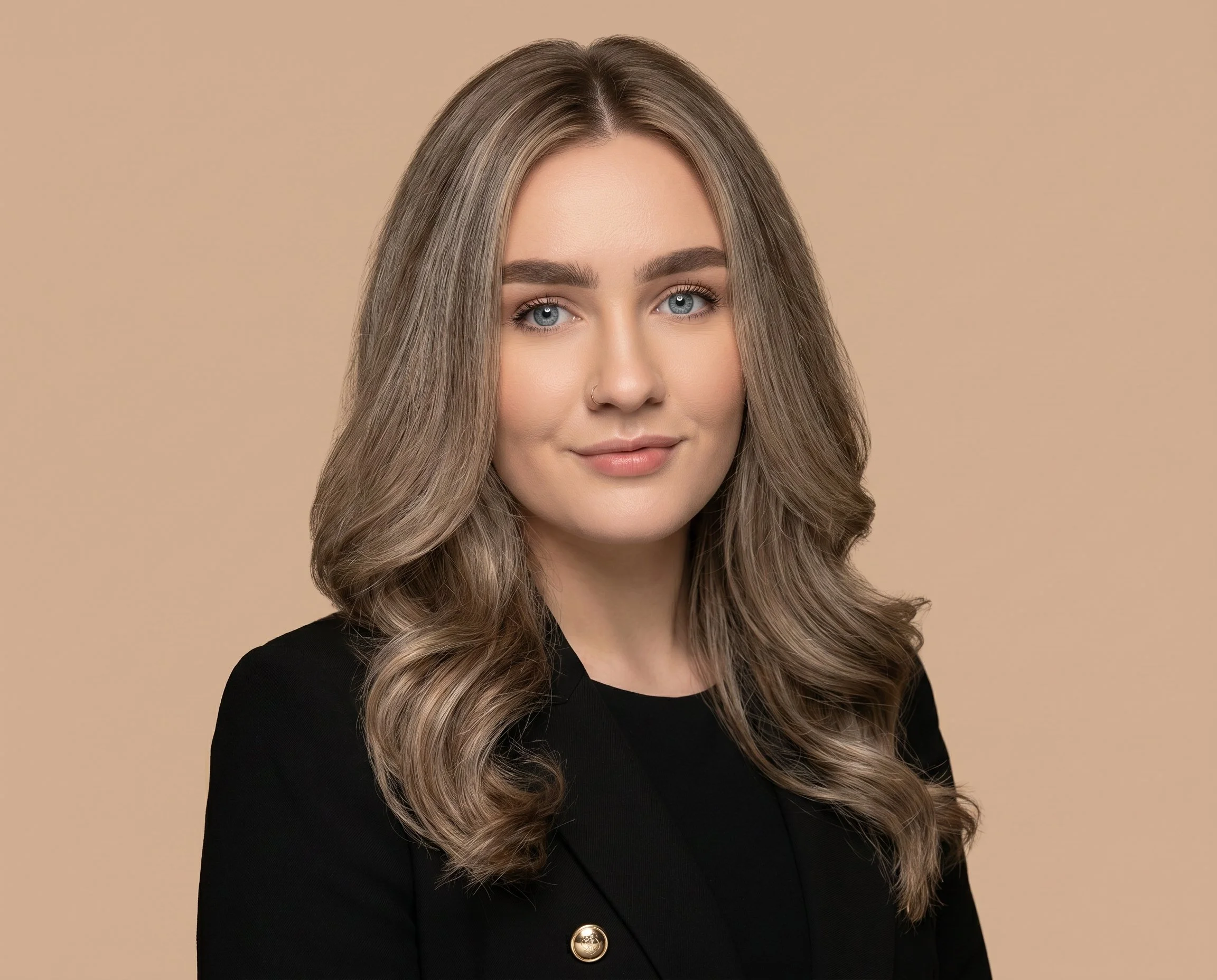 Close-up of a woman with long, wavy, ash blonde hair, light skin, and blue eyes, wearing a black top with a single gold button, against a beige background.