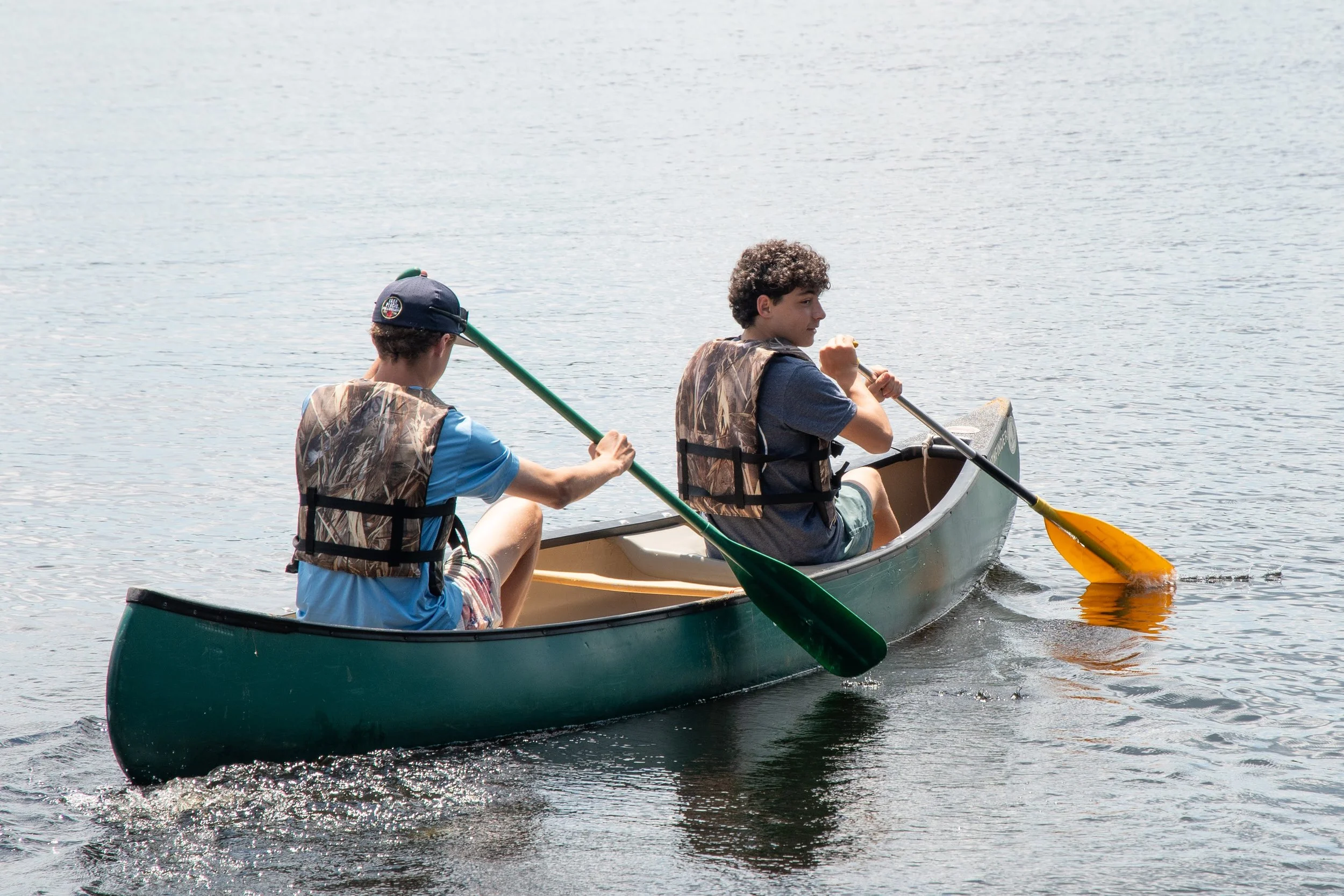 Two boys in a canoe on a lake, paddling with yellow and green paddles. They are wearing life jackets and casual clothes, enjoying a sunny day on the water.