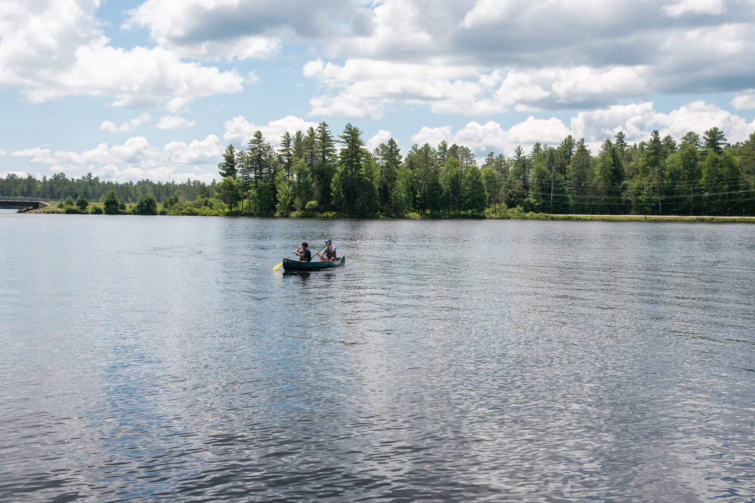 Two people in a canoe paddling on a calm lake surrounded by trees and under a partly cloudy sky.