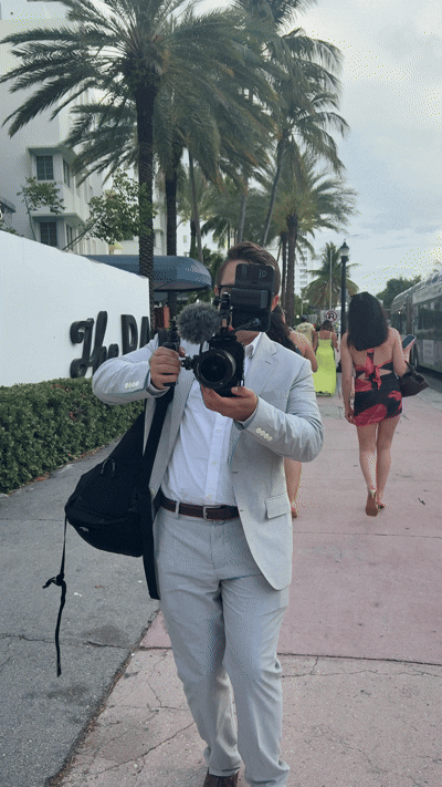 Owen Cappellini is in a light gray suit, holding a camera and facing a mirror, with palm trees and people in the background.