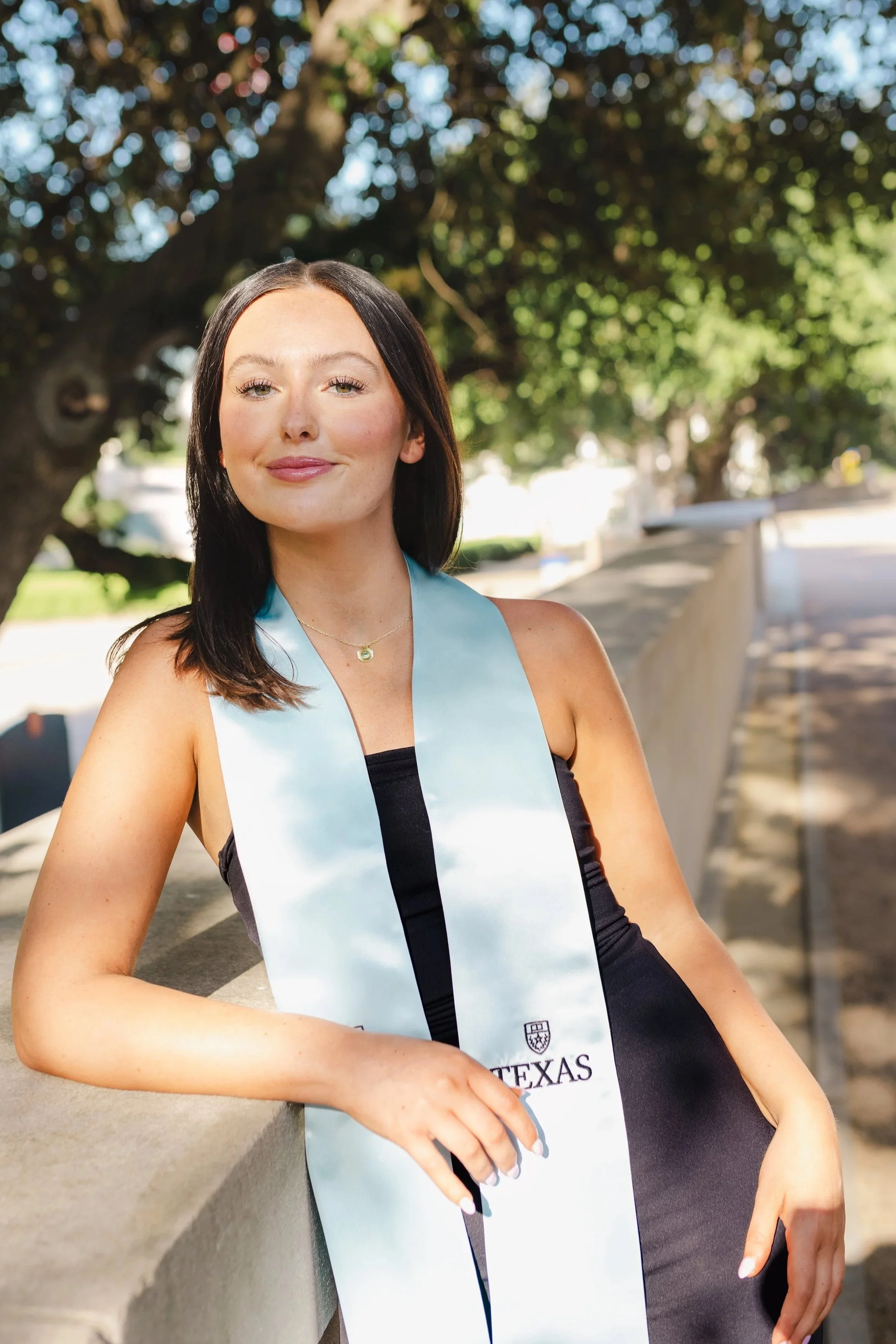 A young woman with dark brown hair smiling outdoors on a sunny day, wearing a black dress and a light blue graduation stole with a Texas emblem.