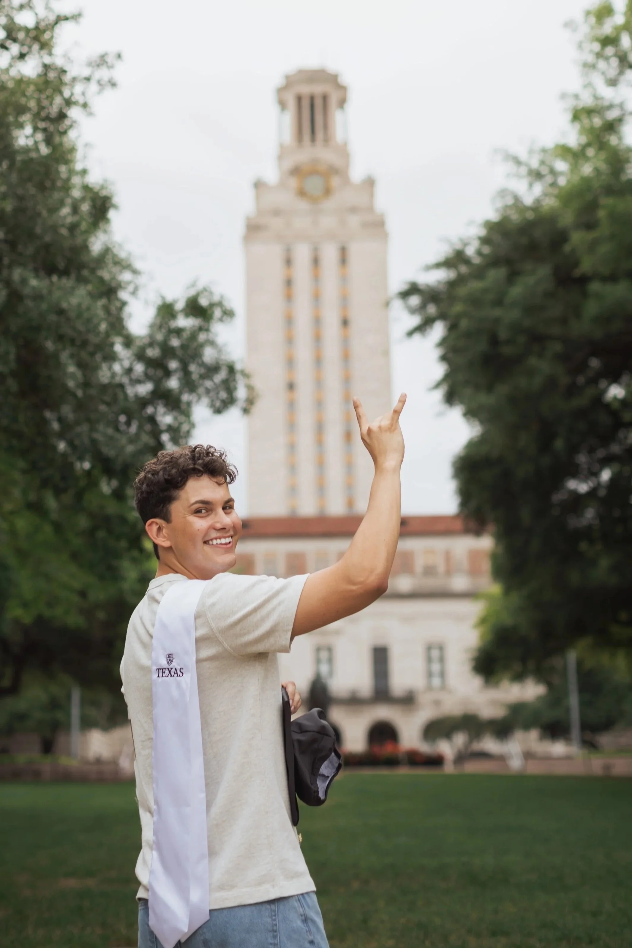 A young man in casual attire, wearing a white graduation stole, throws his graduation cap into the air on a university campus with a tall clock tower in the background.