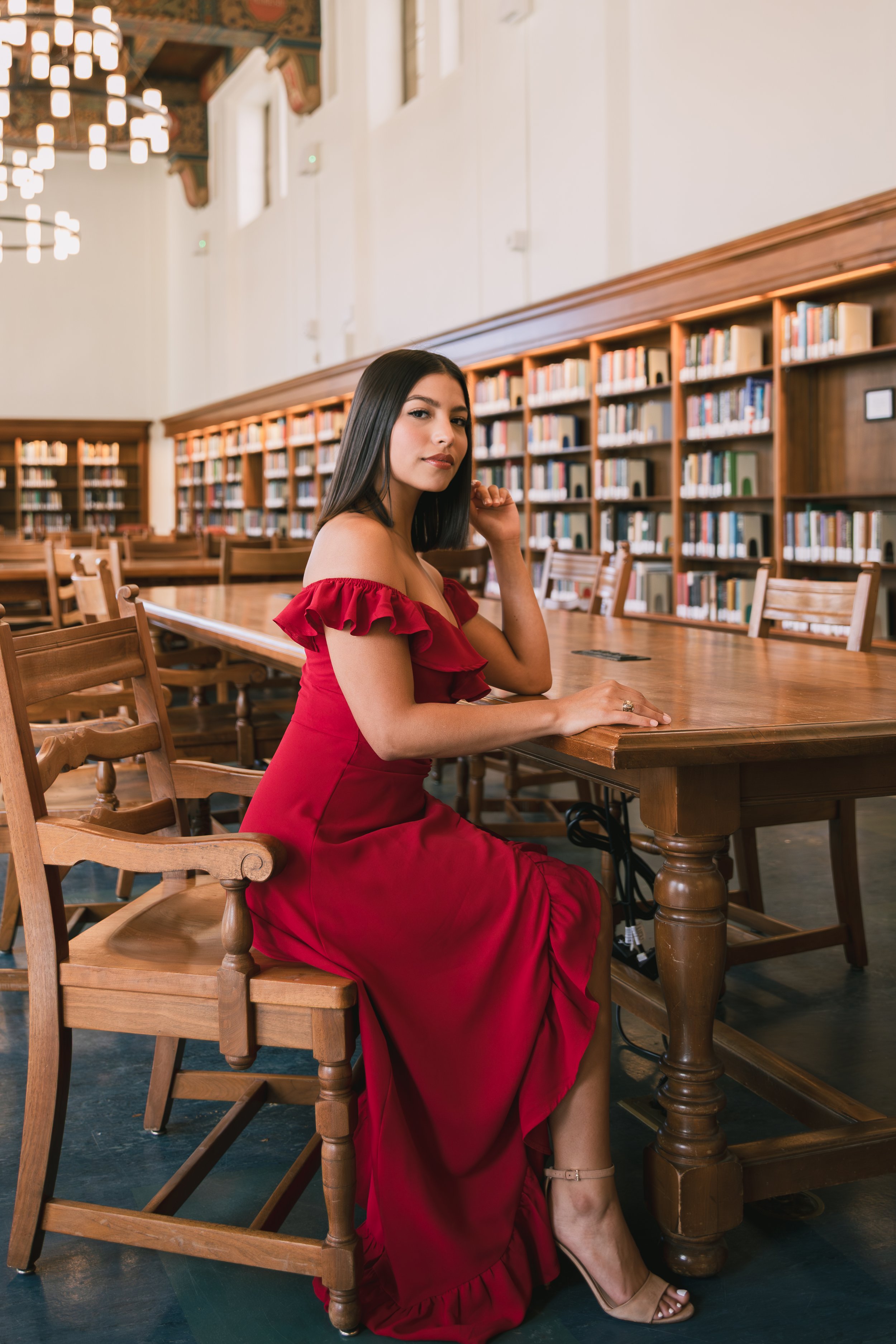A woman in a red off-shoulder dress is sitting at a wooden table in a library with rows of bookshelves in the background.