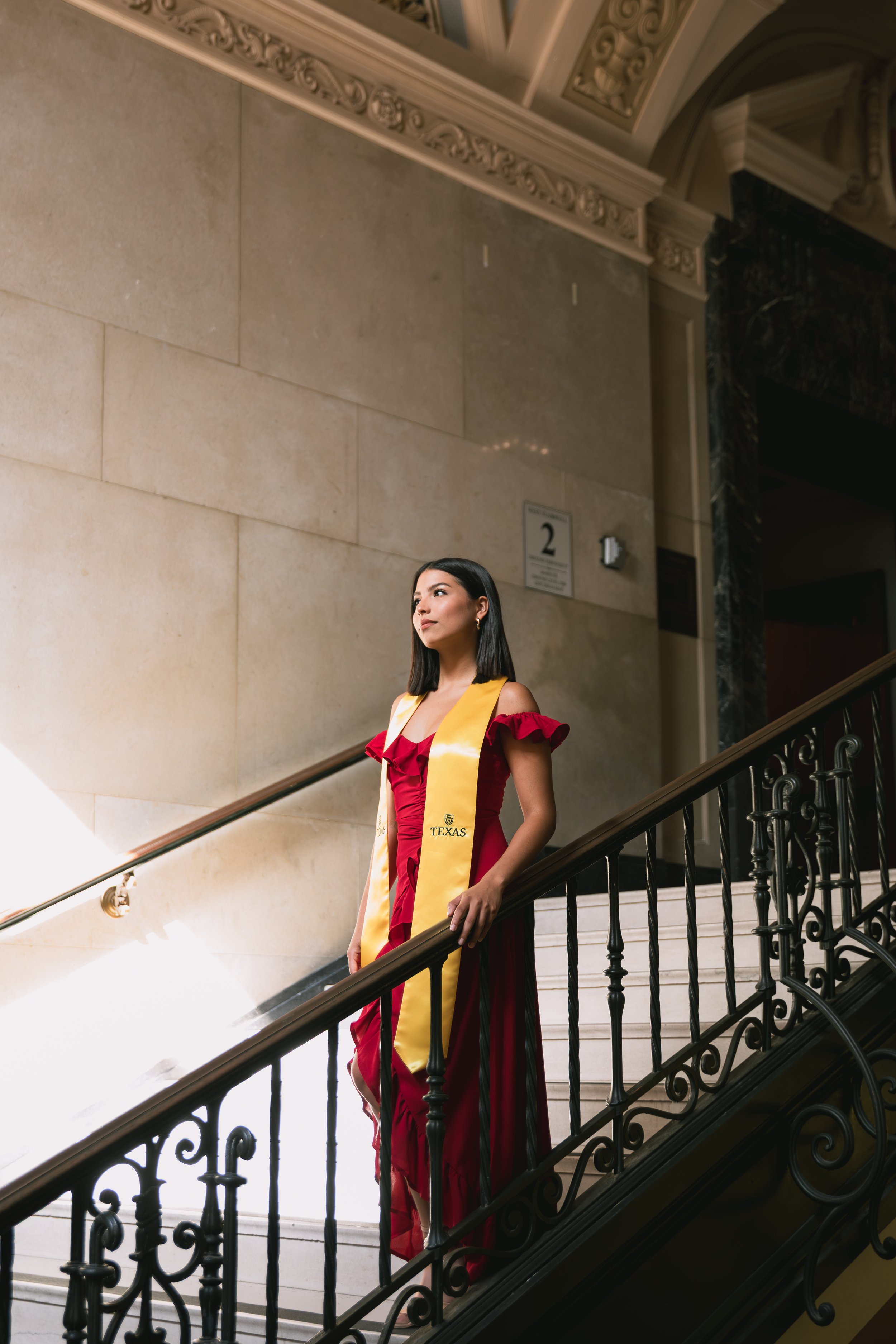 A young woman in an elegant red dress with ruffled sleeves stands on a staircase, wearing a yellow graduation stole with the Texas logo, inside a grand building with ornate architectural details.