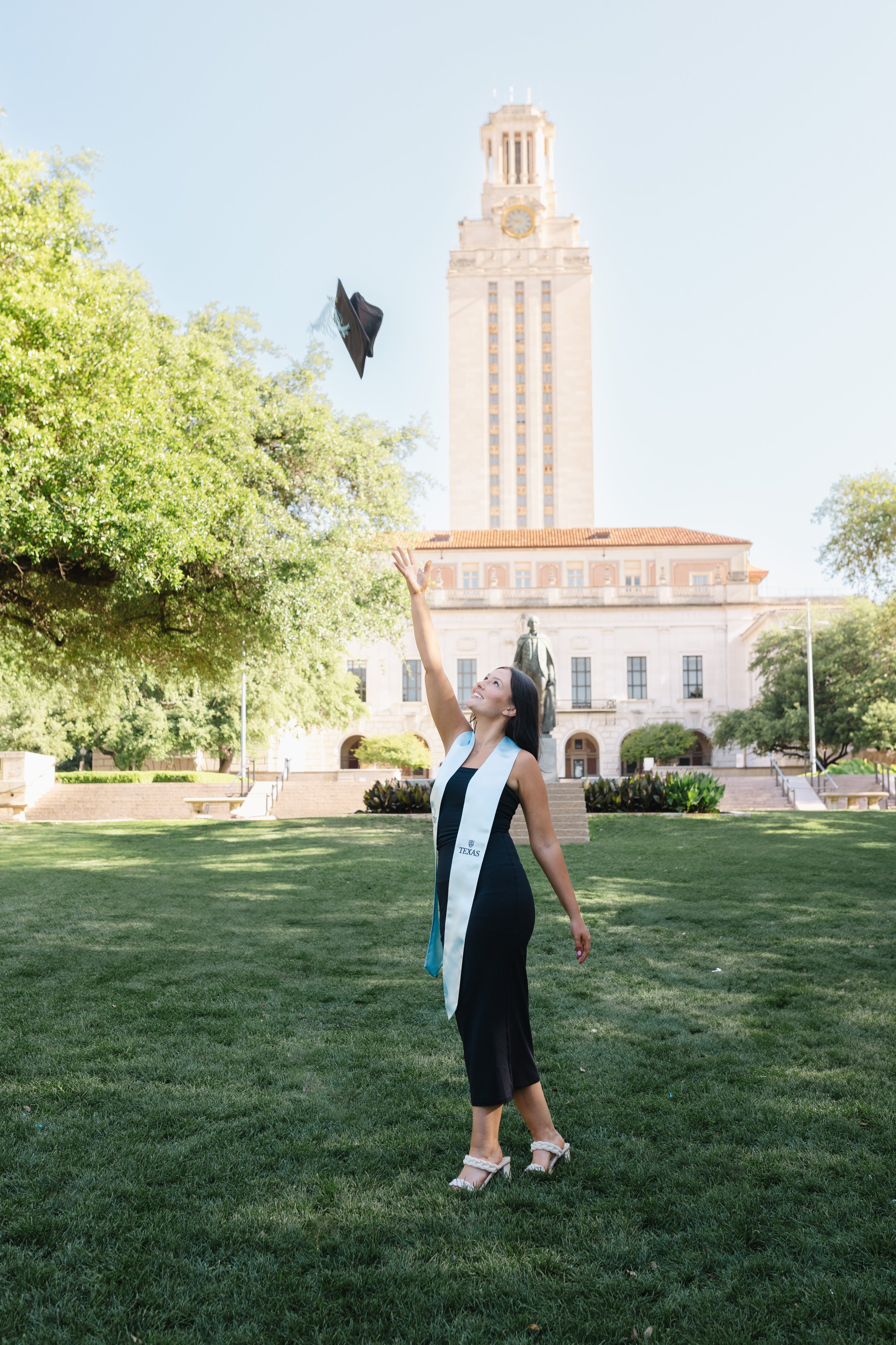 A woman in a black dress and white sash celebrates graduation by tossing her cap in the air on a grassy campus with a tall clock tower and statue in the background.
