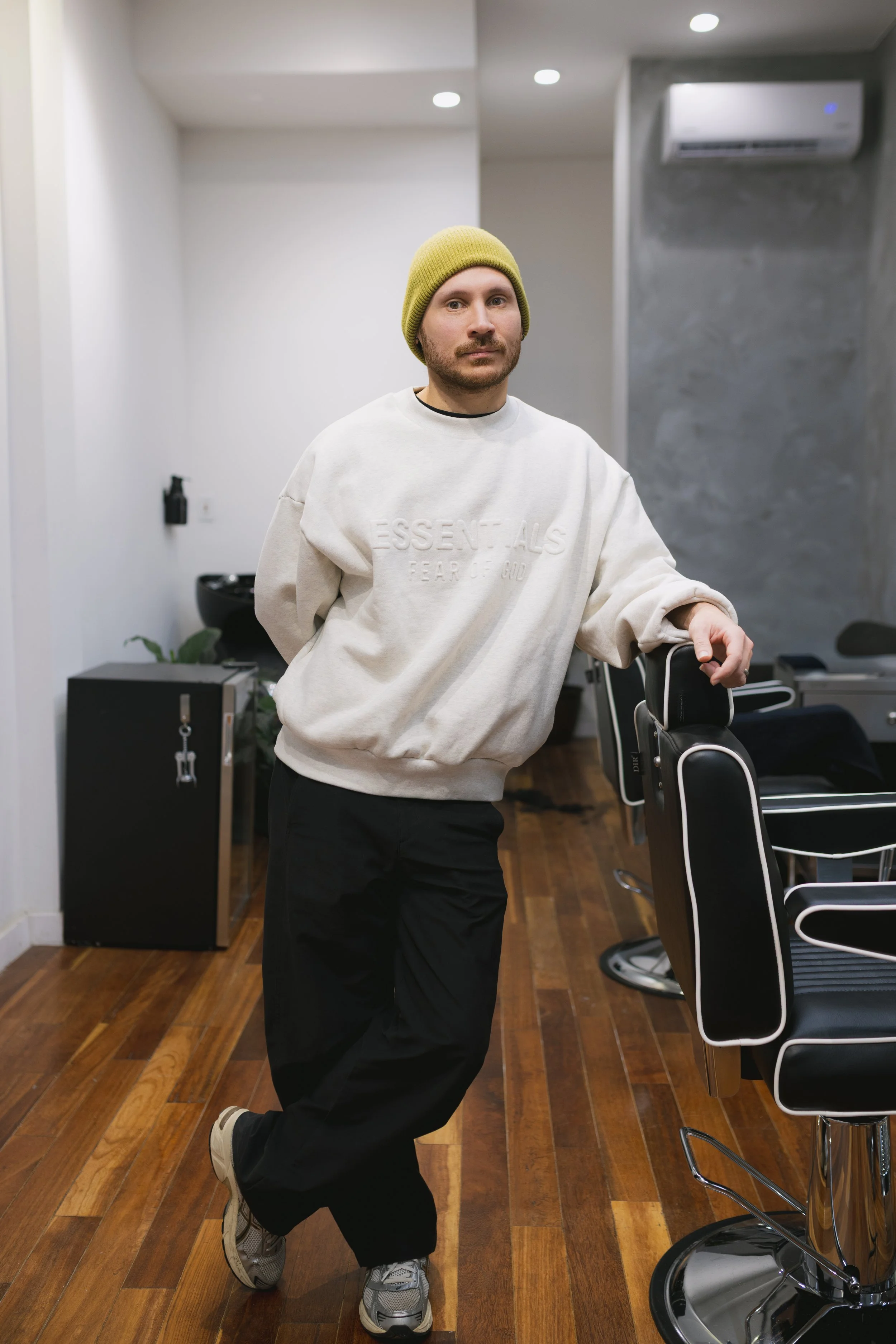 A man wearing a yellow beanie, cream Essentials sweatshirt, black pants, and sneakers leans against a barber chair in a modern barber shop with wooden flooring.