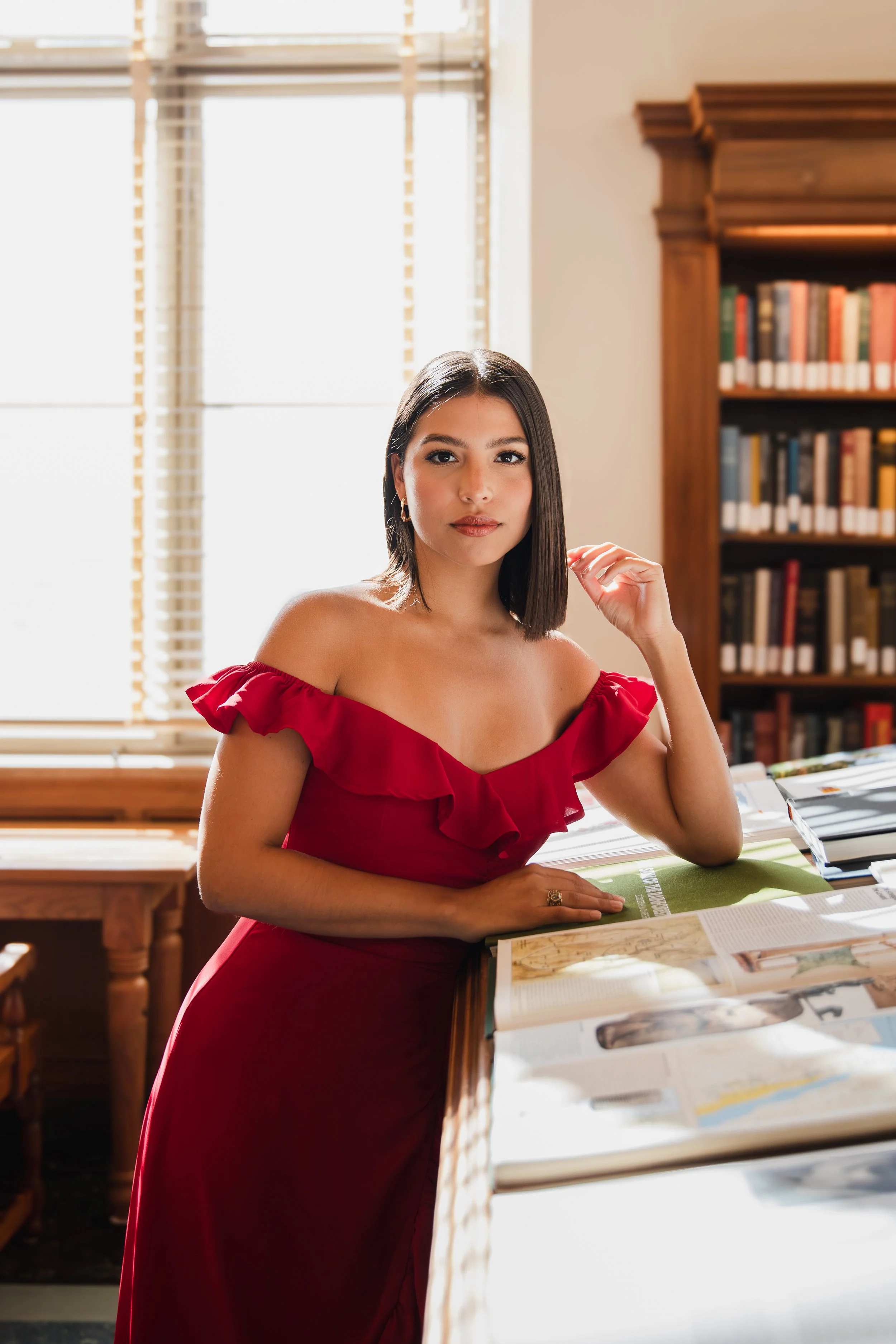 Young woman sitting at a desk in a room with large window and a wooden bookshelf, wearing a red dress.