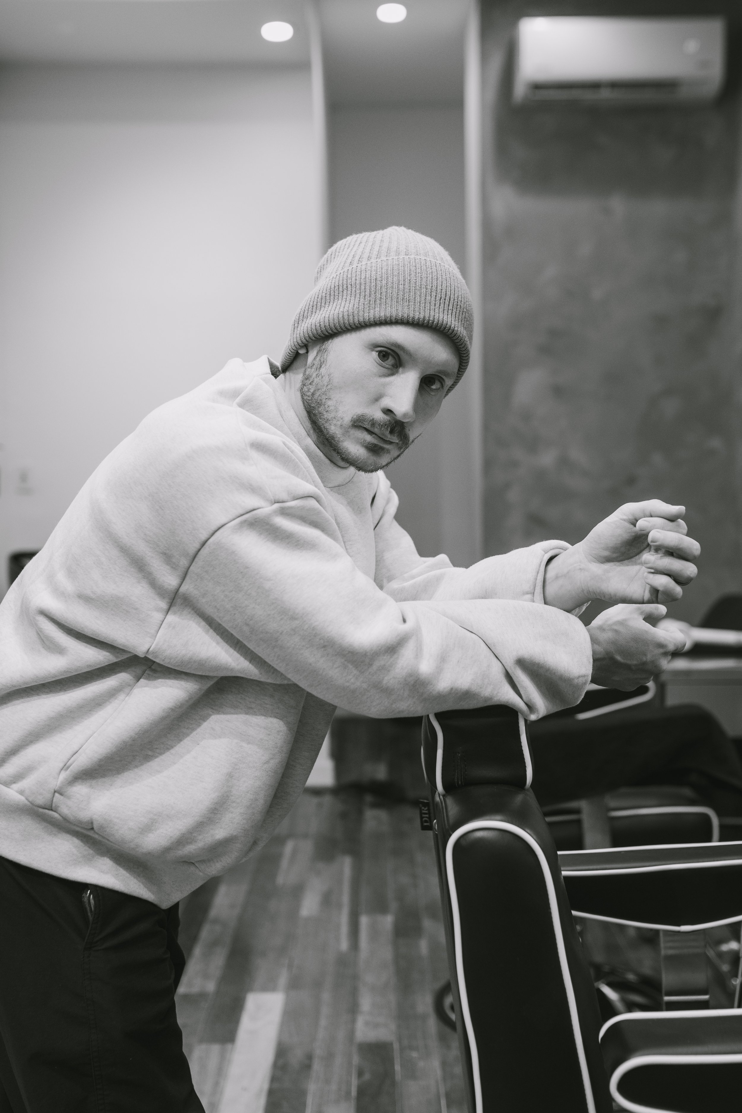 A man wearing a beanie and sweatshirt leaning on a barber chair in a modern barber shop, looking at the camera in an indoor setting.