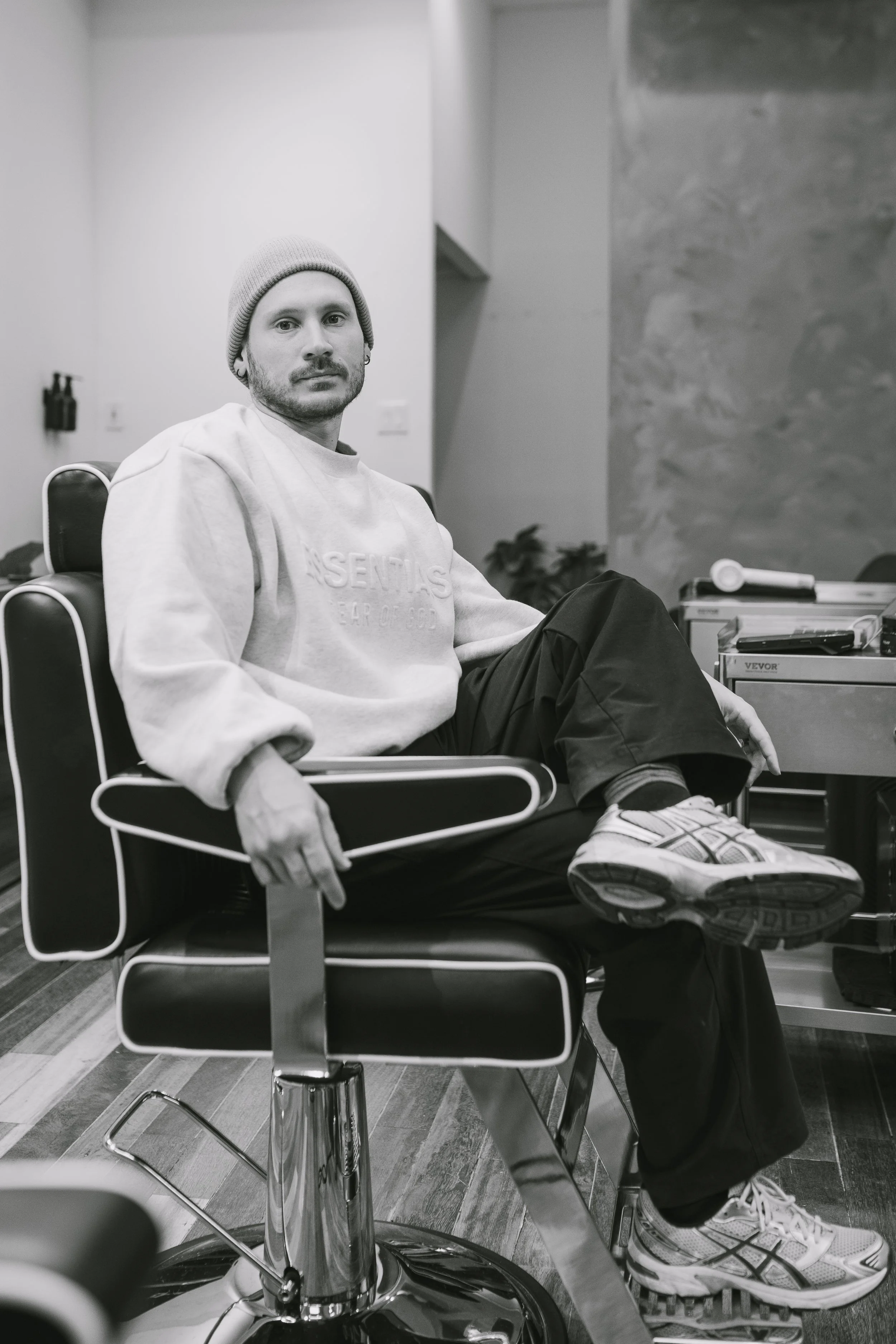 A man with a beanie hat and casual clothing sitting on a barber's chair in a modern salon or barbershop, with barber tools and equipment visible in the background.