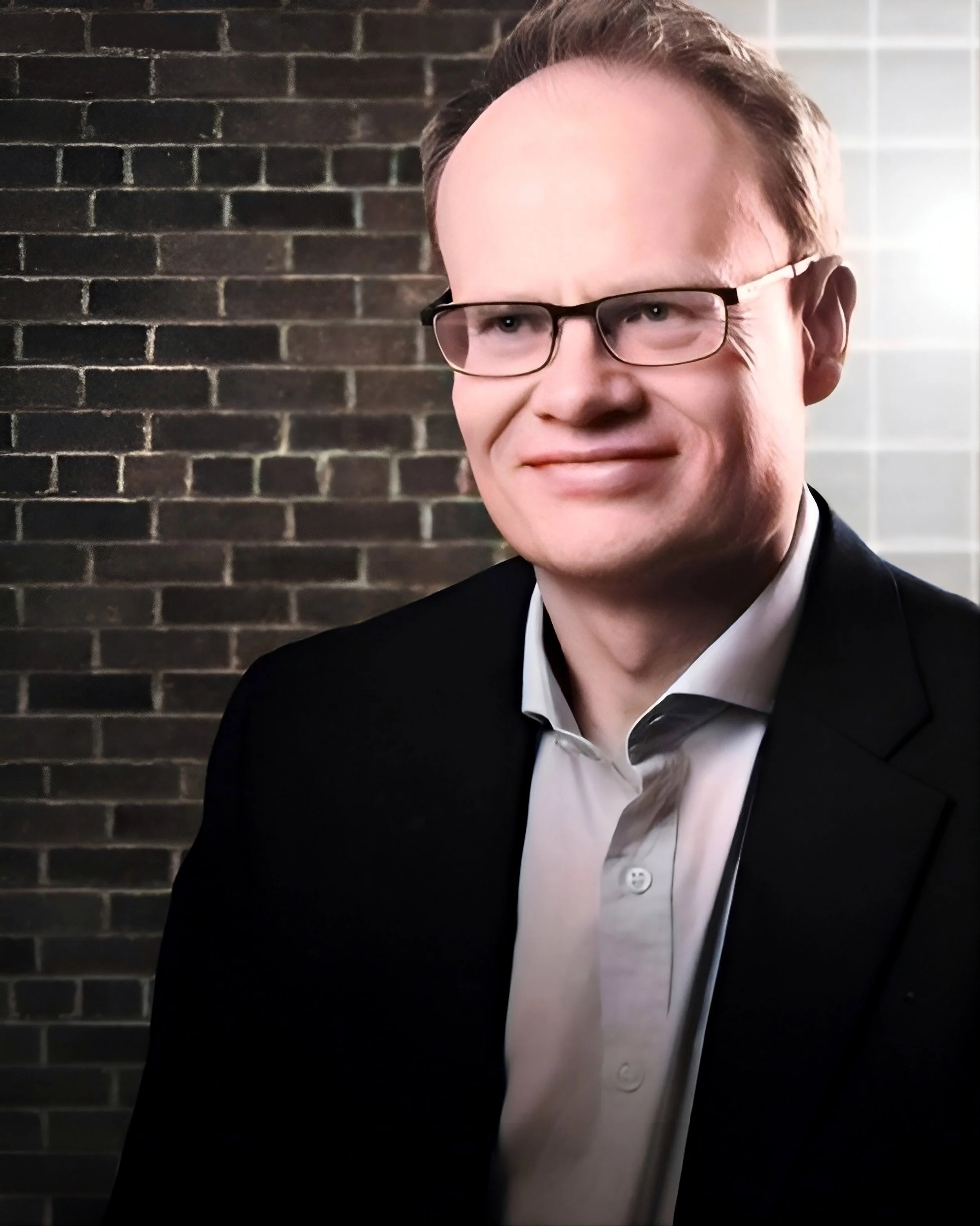 Close-up of a man wearing glasses, a tuxedo, and a white shirt with a black bow tie, standing in front of a brick wall.