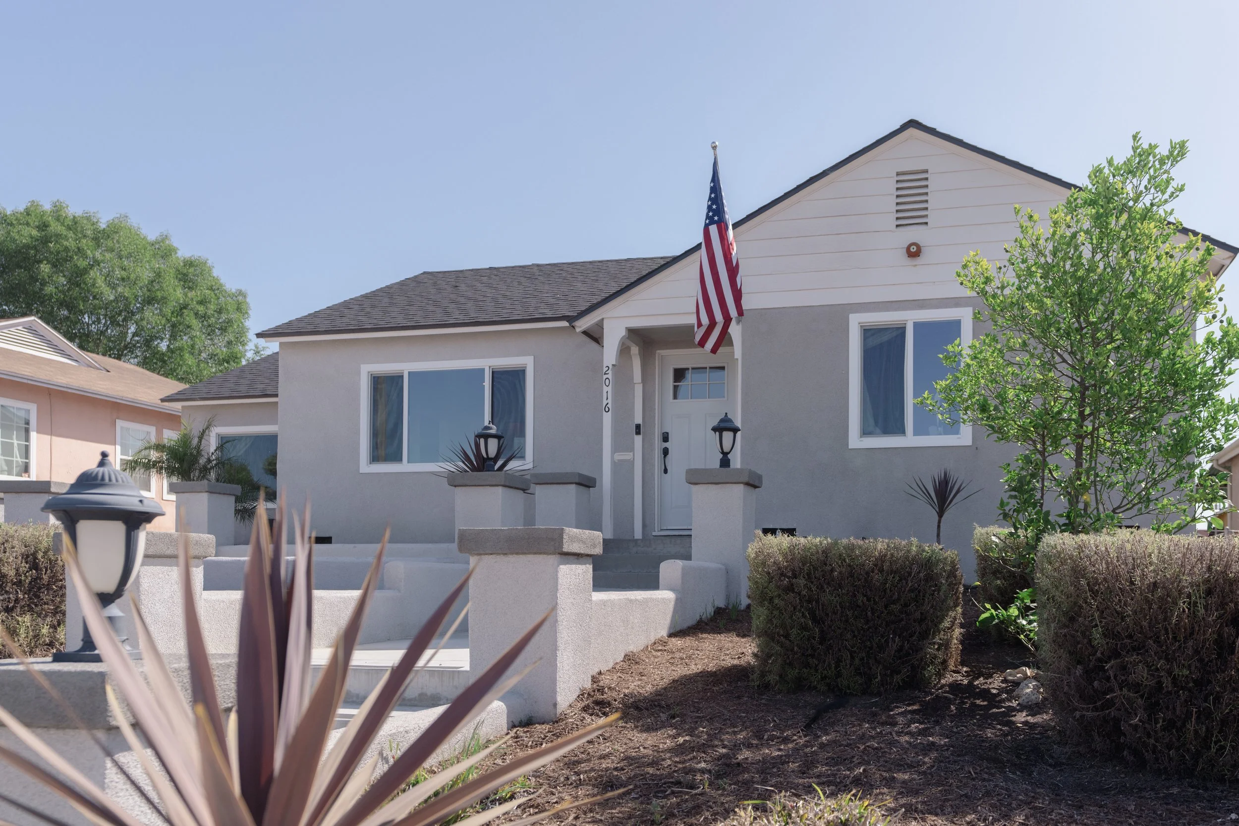A two-story house with a light gray exterior and white trim, American flag displayed on flagpole near the front door, front yard with shrubbery and a small tree, outdoor lamps on pillars, neighboring houses in background, and a clear blue sky.