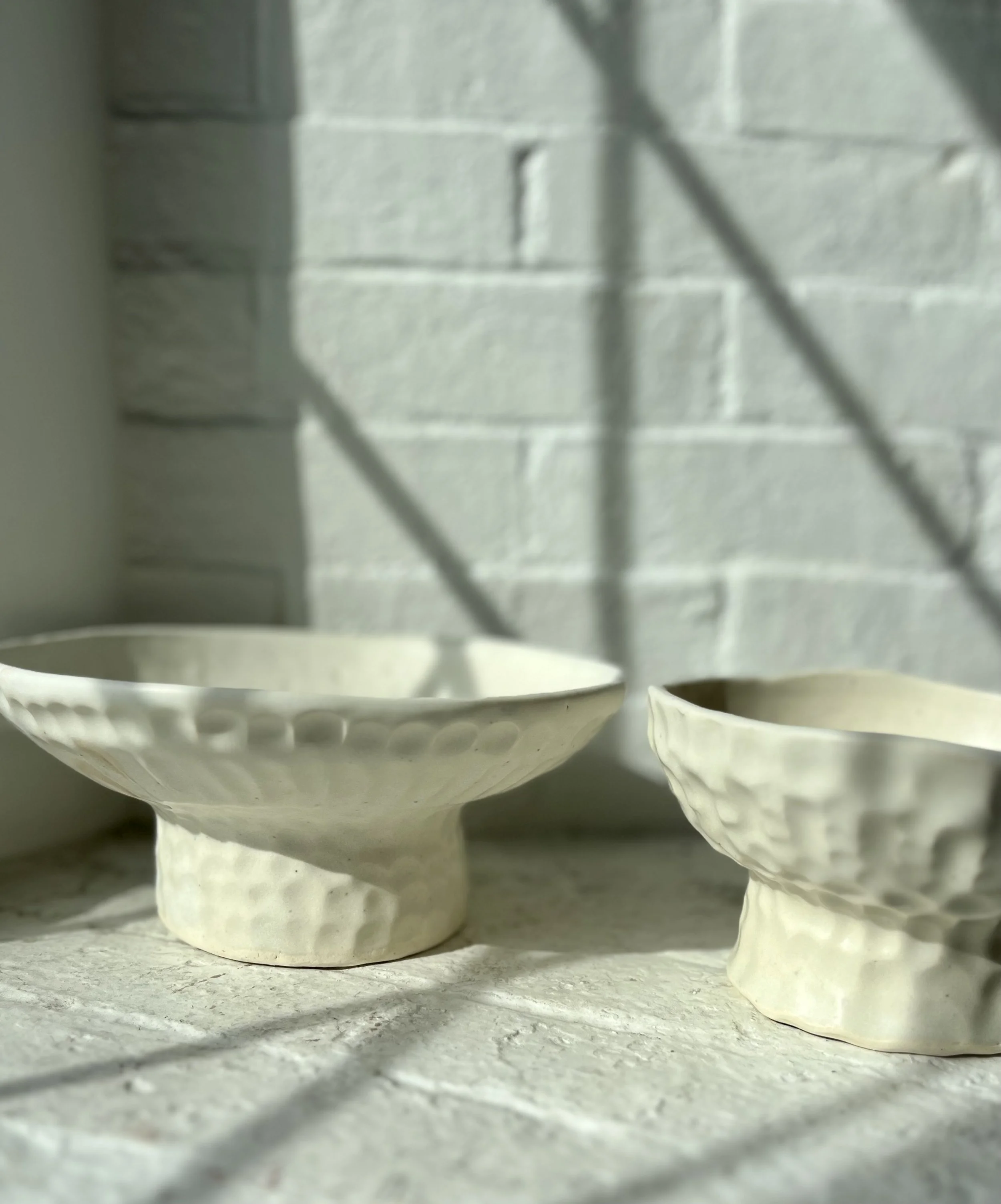 Two white ceramic bowls with textured surfaces sitting on a light-colored surface, with soft sunlight and shadows cast on a white brick wall background.