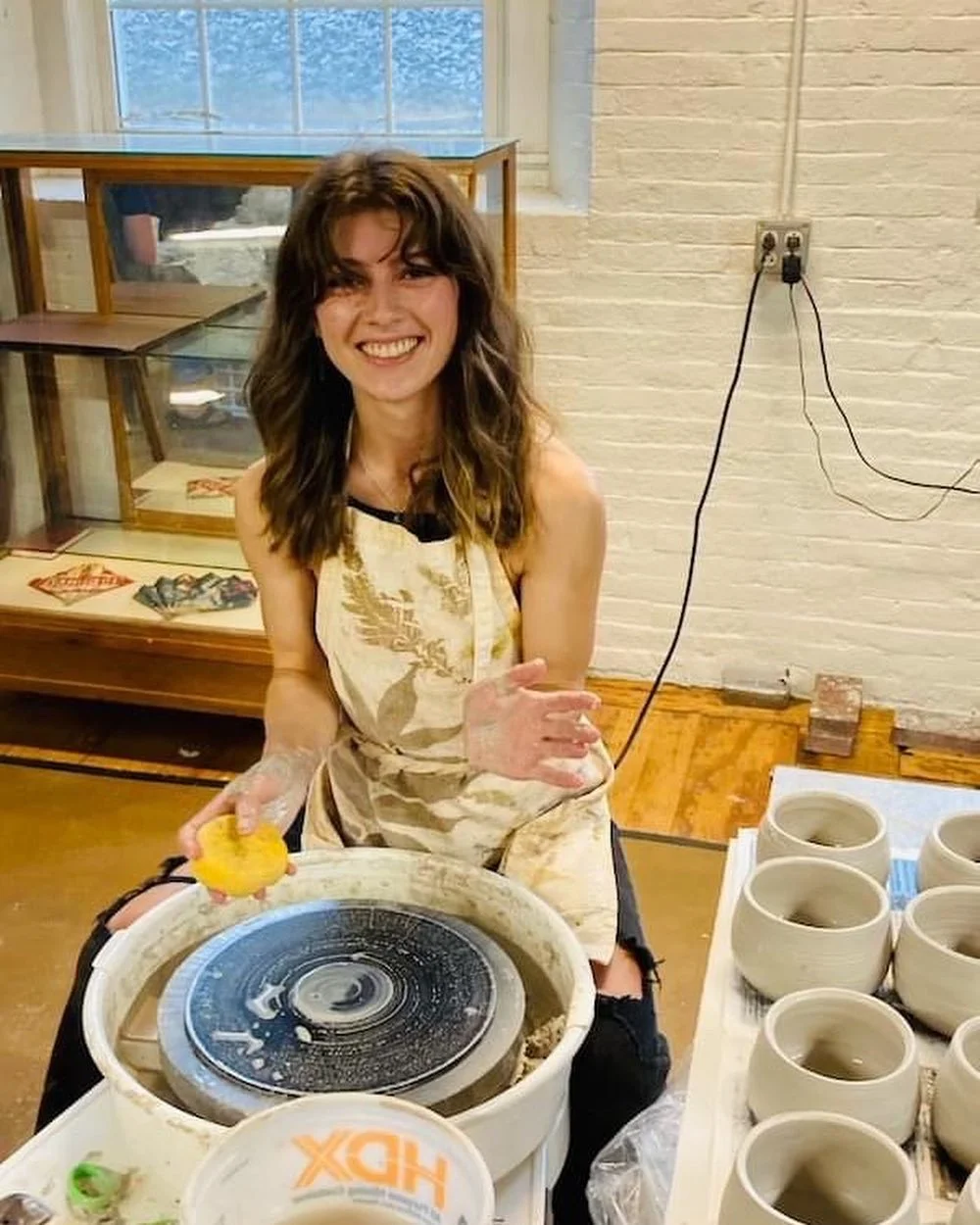 A young woman with wavy brown hair and a sleeveless top sits at a pottery wheel, smiling and waving at the camera, while making a ceramic piece in a studio with shelves of pottery and a brick wall in the background.