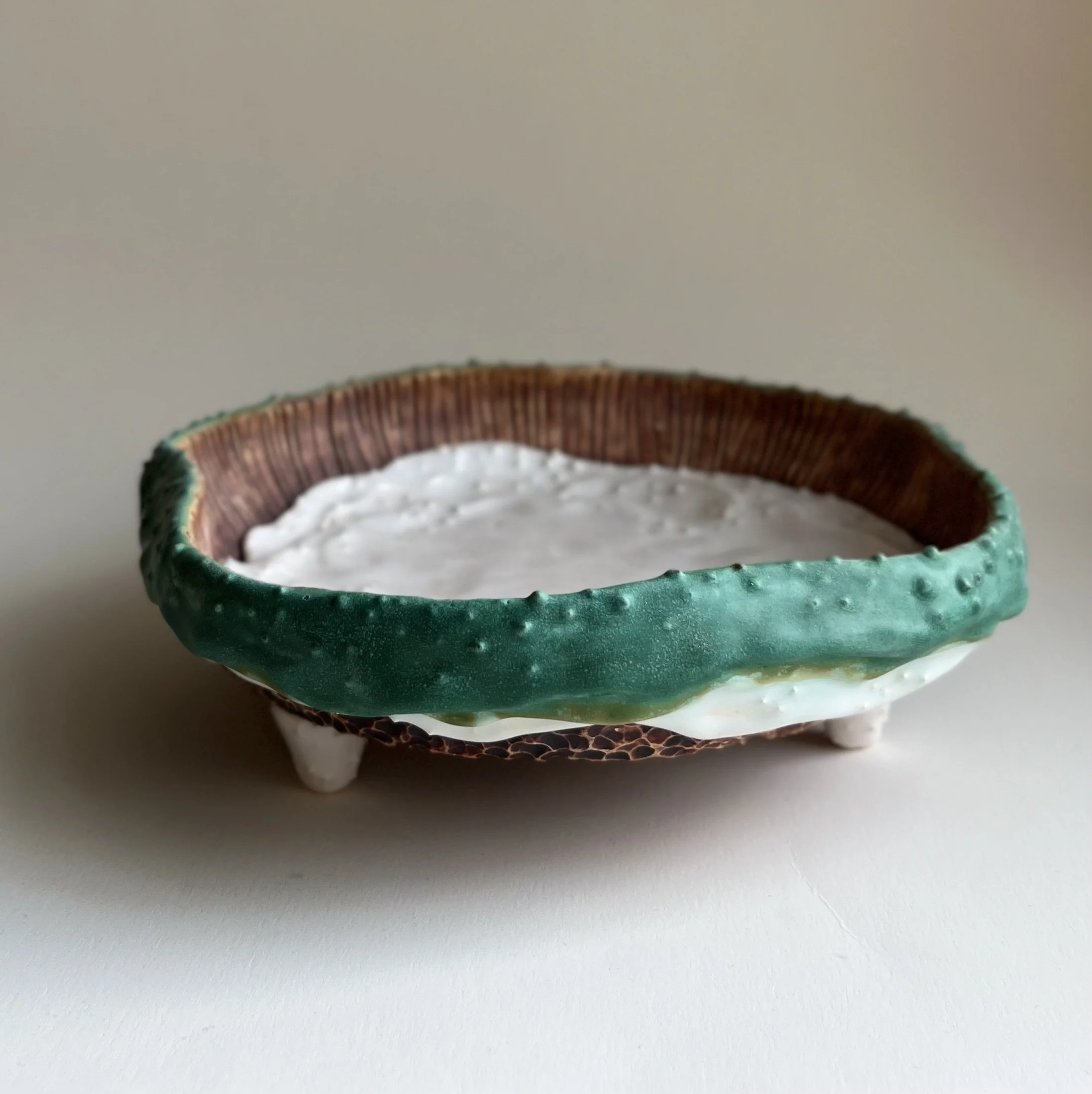 Ceramic bowl with green and brown exterior and white interior, standing on three small legs on a white surface.