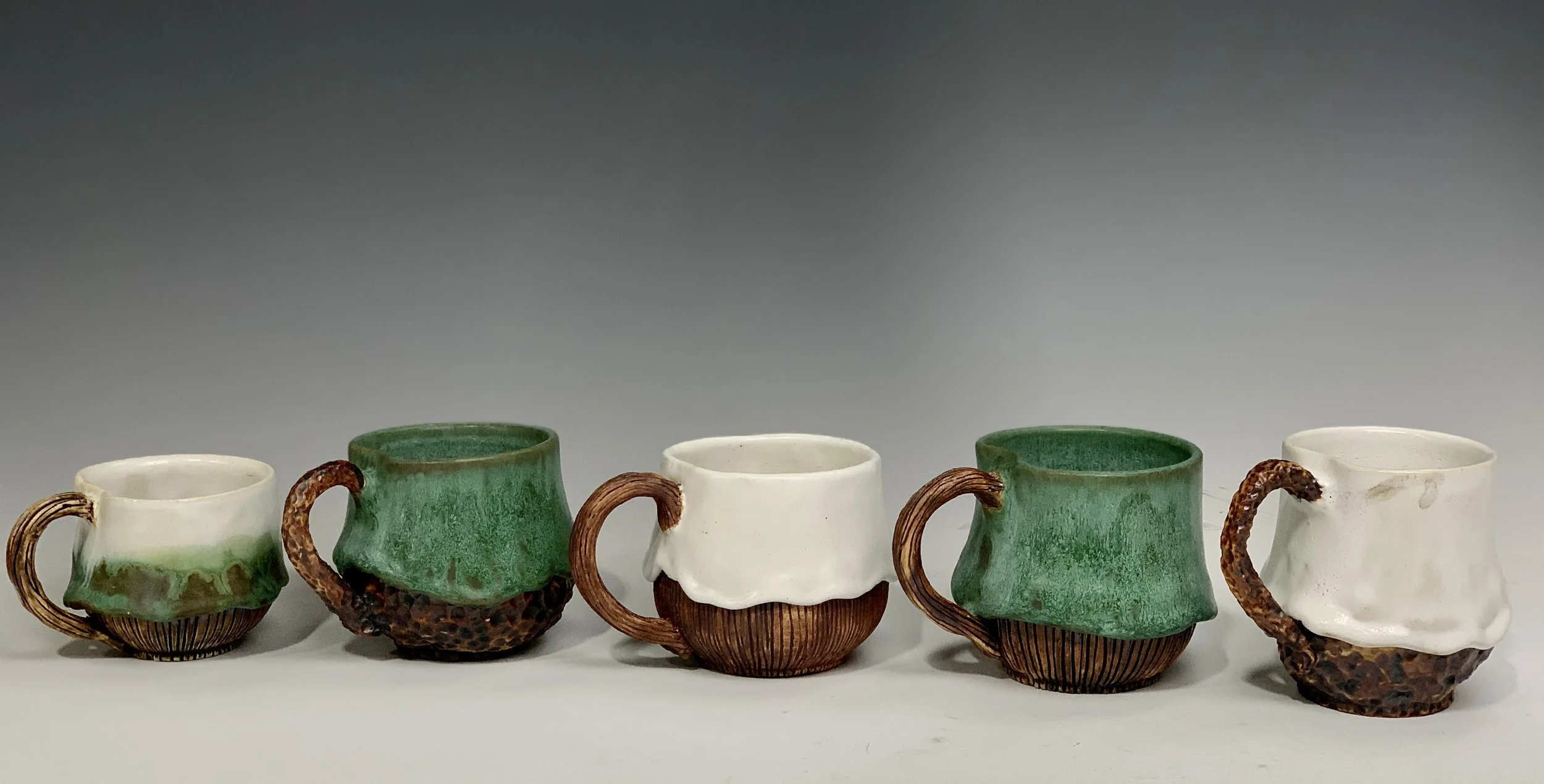 Set of five ceramic coffee cups with varying green, white, and brown glaze, featuring rustic handles and textured bases, arranged in a row on a plain background.