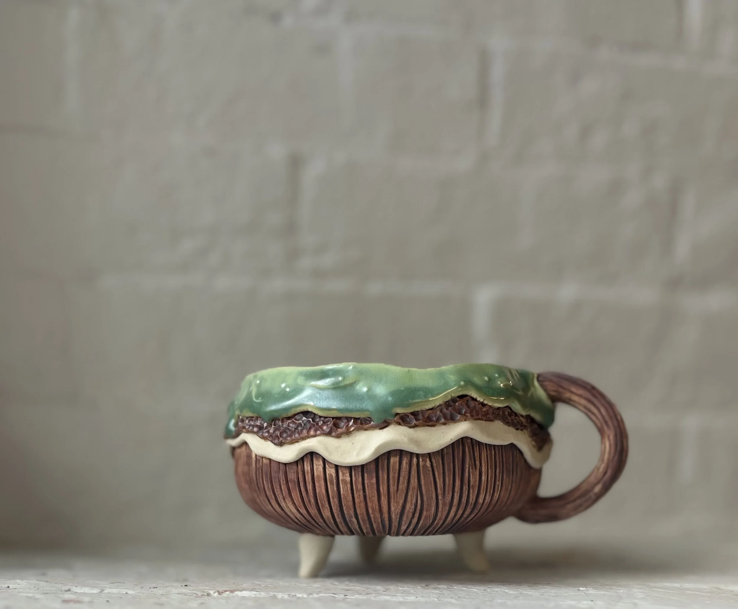 A small decorative ceramic teacup with a textured brown exterior and a curved handle, decorated with realistic-looking green, white, and brown glaze resembling a layered dessert. The cup is placed on a white surface against a light gray brick wall ba