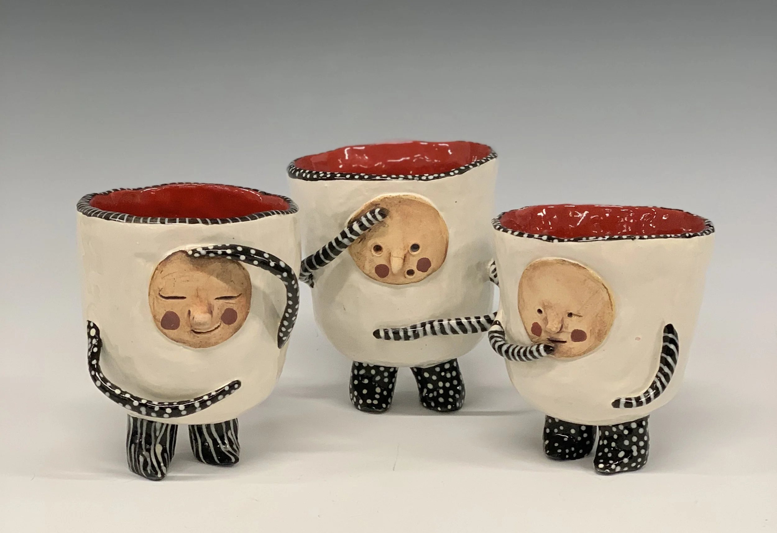 Three whimsical ceramic cups with faces, arms, and legs, decorated in black and white polka dots, red interior, and beige exterior with face designs, standing on a white surface.