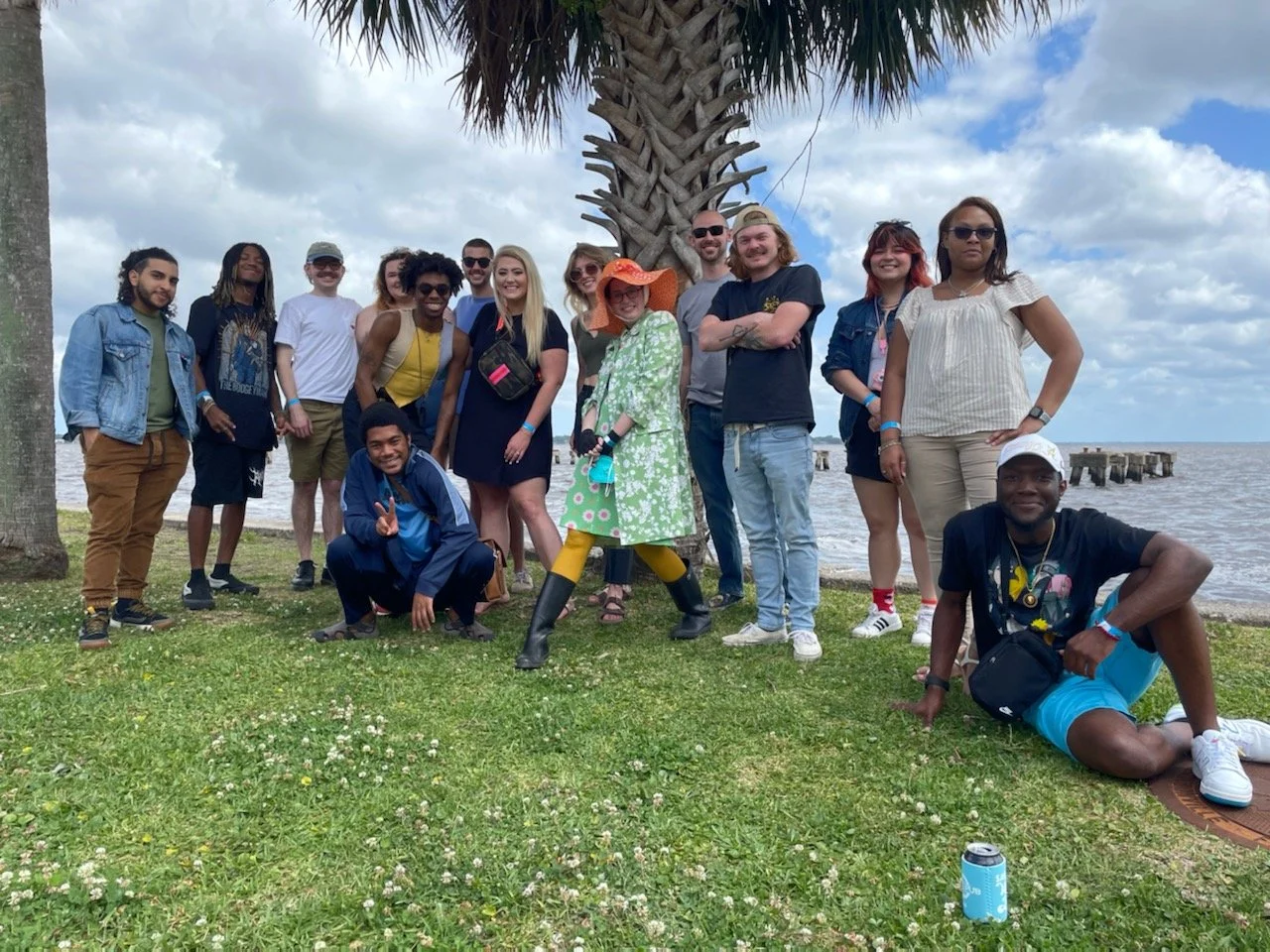 Group of people posing outdoors near the water with a palm tree, cloudy sky, and pier in the background.
