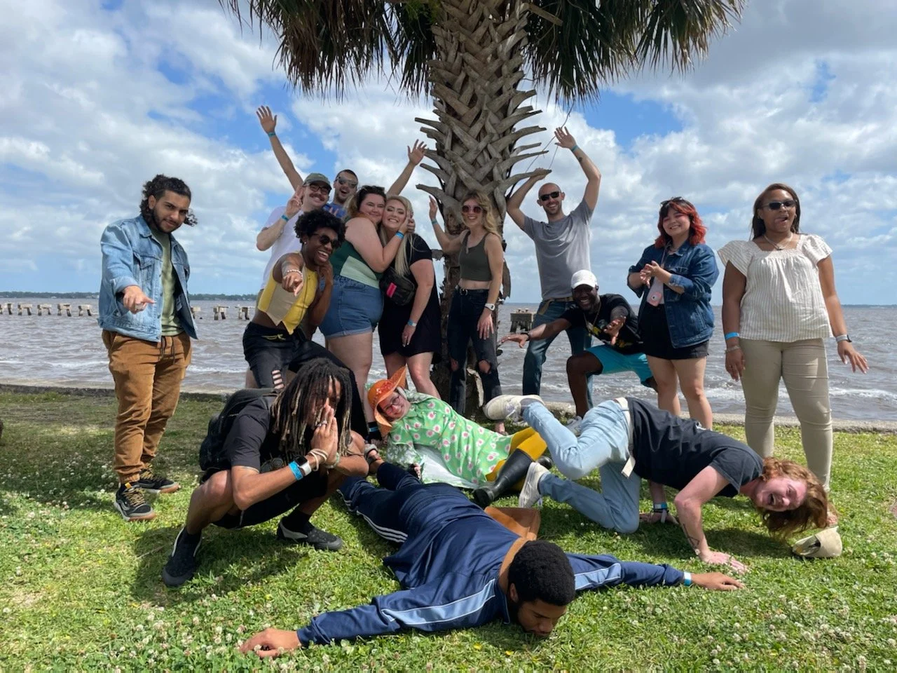 A group of diverse people posing and laughing outdoors near a palm tree by the water under a cloudy sky.